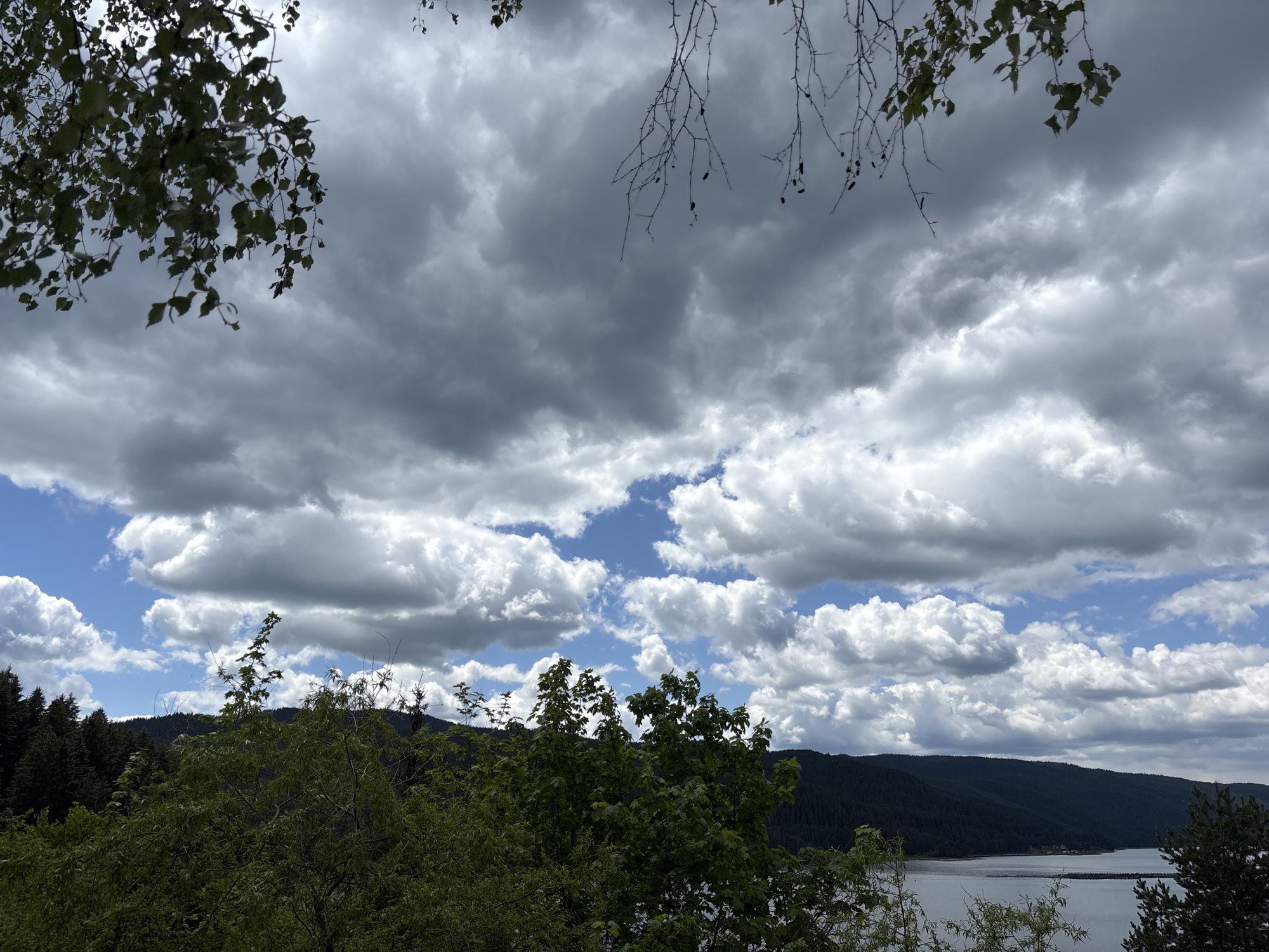 The image presents a landscape in Dospat, Bulgaria, featuring an overcast sky with cumulonimbus clouds interspersed with patches of blue. In the foreground, green foliage frames the view, obscuring the lower portion of the scene. A body of water, likely a reservoir or lake, is visible in the middle ground, with forested hills stretching along the horizon. The atmosphere suggests a day with mixed weather conditions.