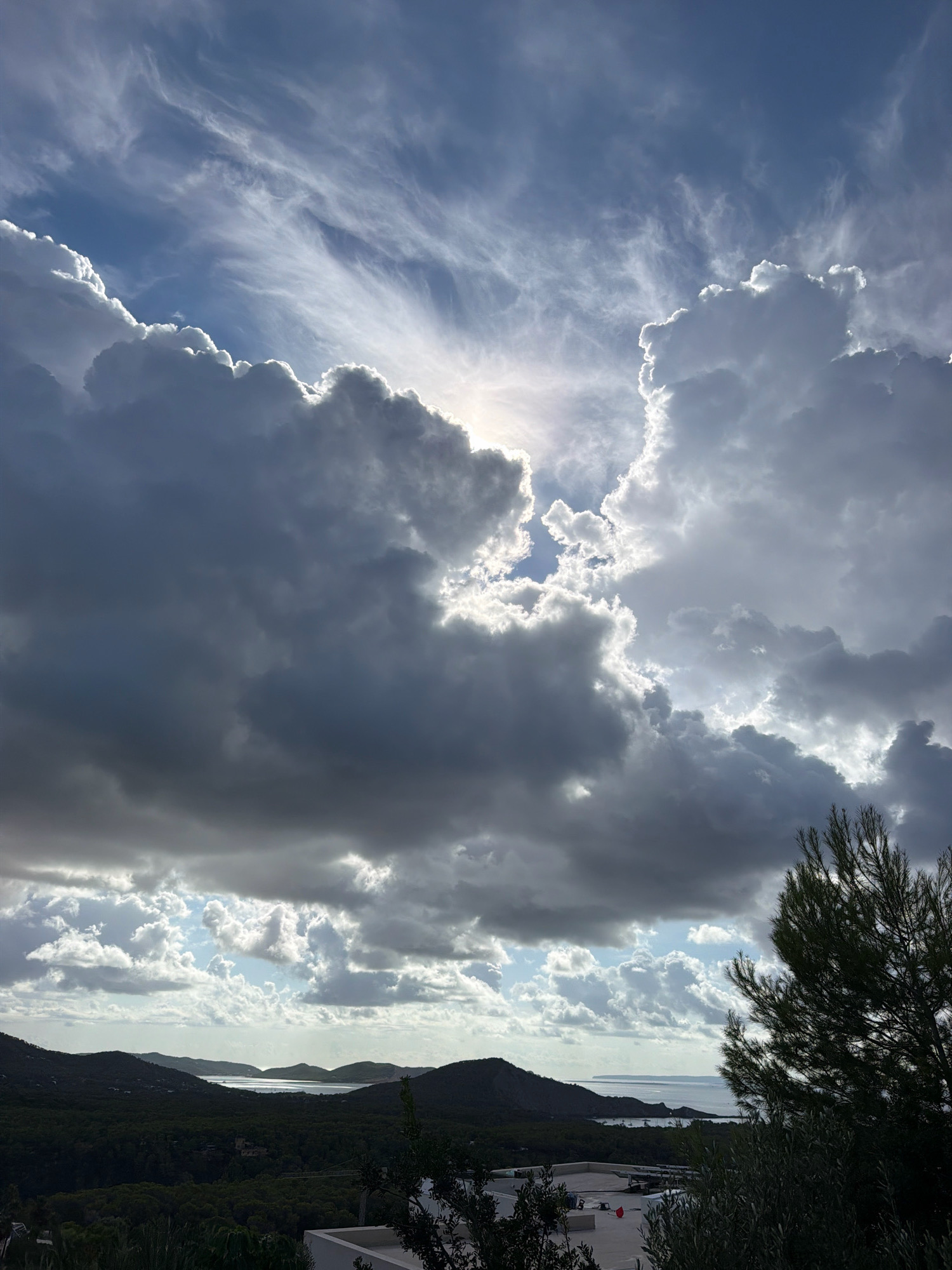 This is a vertical, high-angle shot of a cloudy sky over a coastal landscape in Sant Josep de sa Talaia, Spain. The sun is partially obscured by dramatic, dark grey clouds in the foreground, which are backlit by the sun, creating a bright halo effect around their edges. Wispy, white clouds stretch across the blue sky in the upper portion of the frame. Below the clouds, rolling, dark green hills covered in trees lead down to a body of water. In the distance, islands are visible on the horizon. A portion of a modern white building and a tall, dark green pine tree with sparse needles are in the lower right corner of the image, framing the scene.