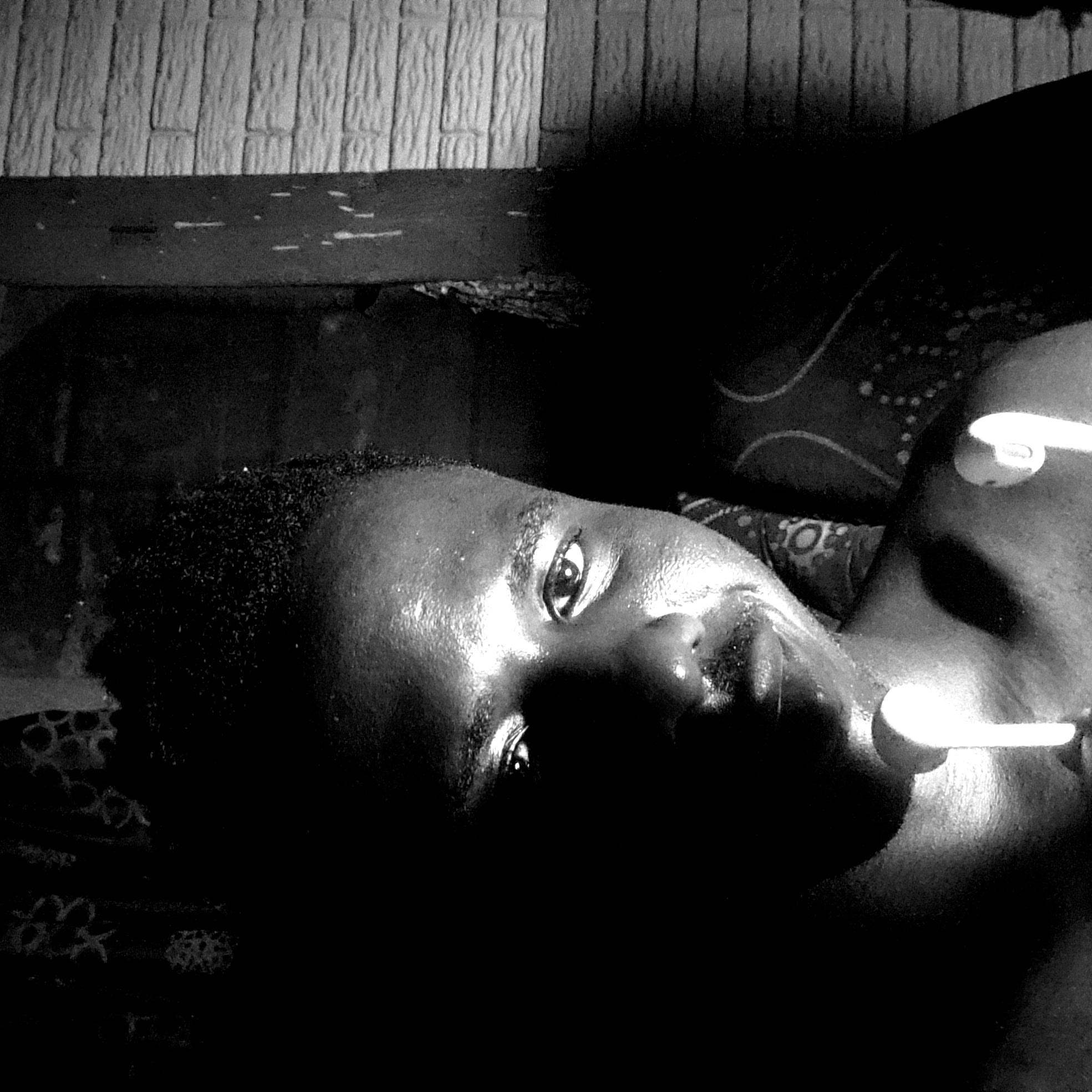 A black and white photograph shows a person lying down, possibly on a bed with patterned sheets. The person appears to be wearing earbuds. The environment is dimly lit. Location of Narayi, Nigeria cannot be verified from provided image.