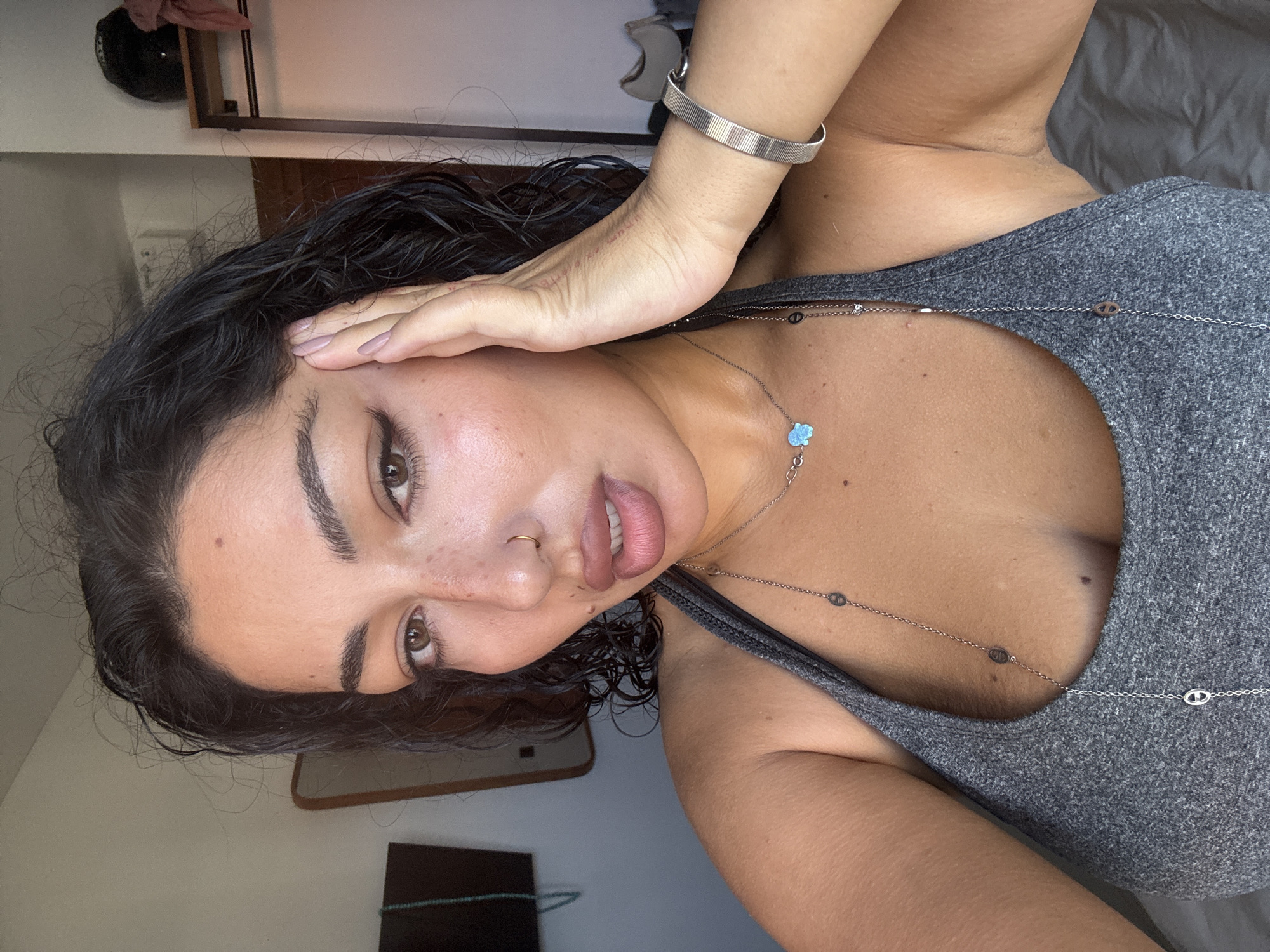 A young woman with dark hair is posing for a selfie. She is wearing a grey tank top and several pieces of jewelry, including a necklace with a small blue pendant and a silver bracelet. Her hand is resting on her face. The background appears to be an indoor setting, possibly a bedroom, with a mirror and a clothing rack visible. There are no obvious indicators of the specific location within Kuta Selatan, Indonesia, and the provided image does not offer enough context to determine this. The lighting suggests it is daytime.