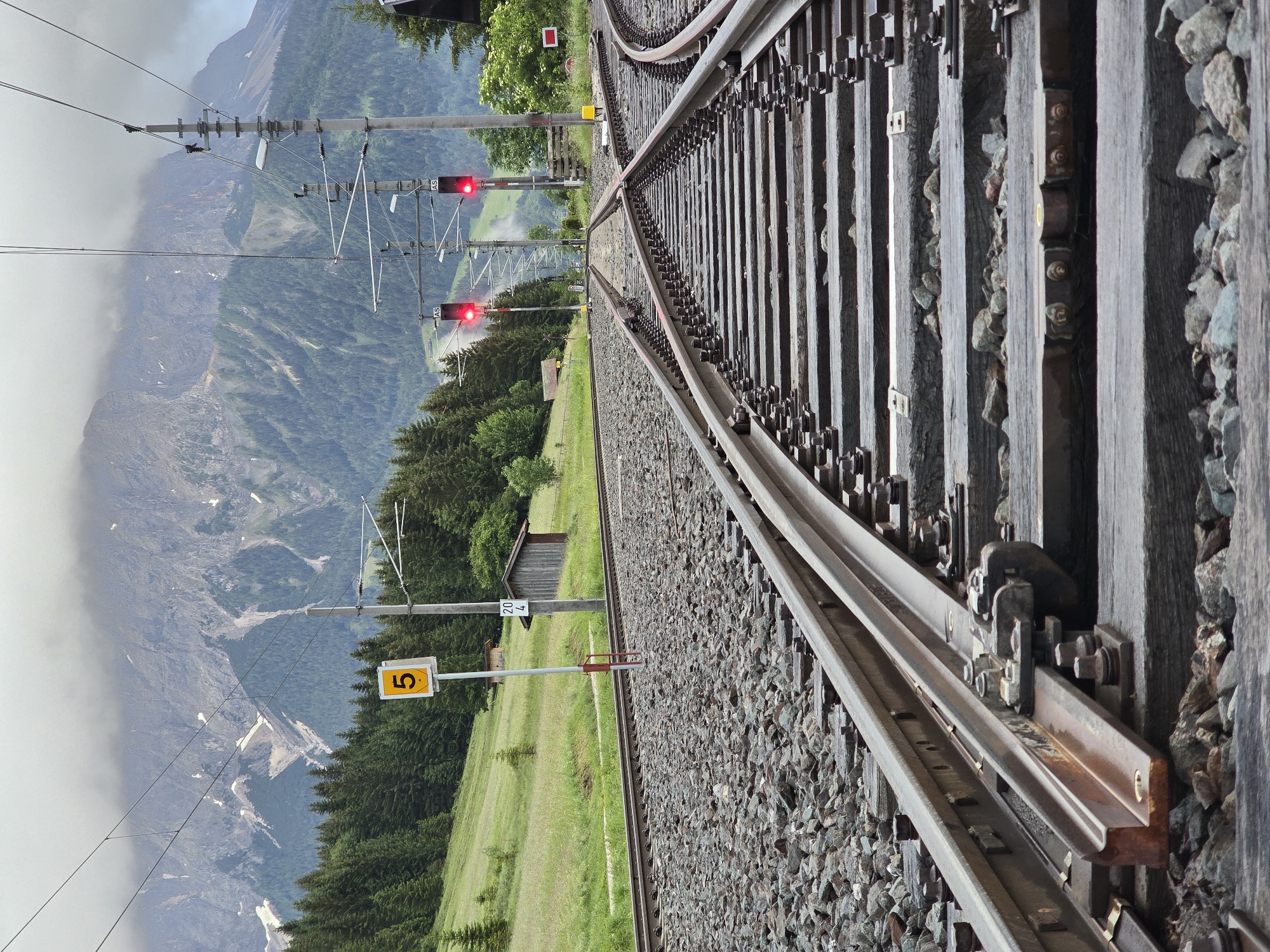 The image depicts a railway track junction in Lermoos, Austria, with a steep, forested mountainside and a cloudy sky in the background. The railway tracks are made of steel rails and wooden sleepers, with ballast stones filling the spaces between them. A sign with the number "5" is visible on the left side of the tracks. Two railway signals displaying red lights are positioned further down the tracks, indicating a stop. Two small wooden sheds are situated on the grassy embankment to the right of the tracks. The scene appears to be a natural landscape setting, with no people or animals visible. The overall impression is one of a functional railway infrastructure integrated into a scenic mountain environment.