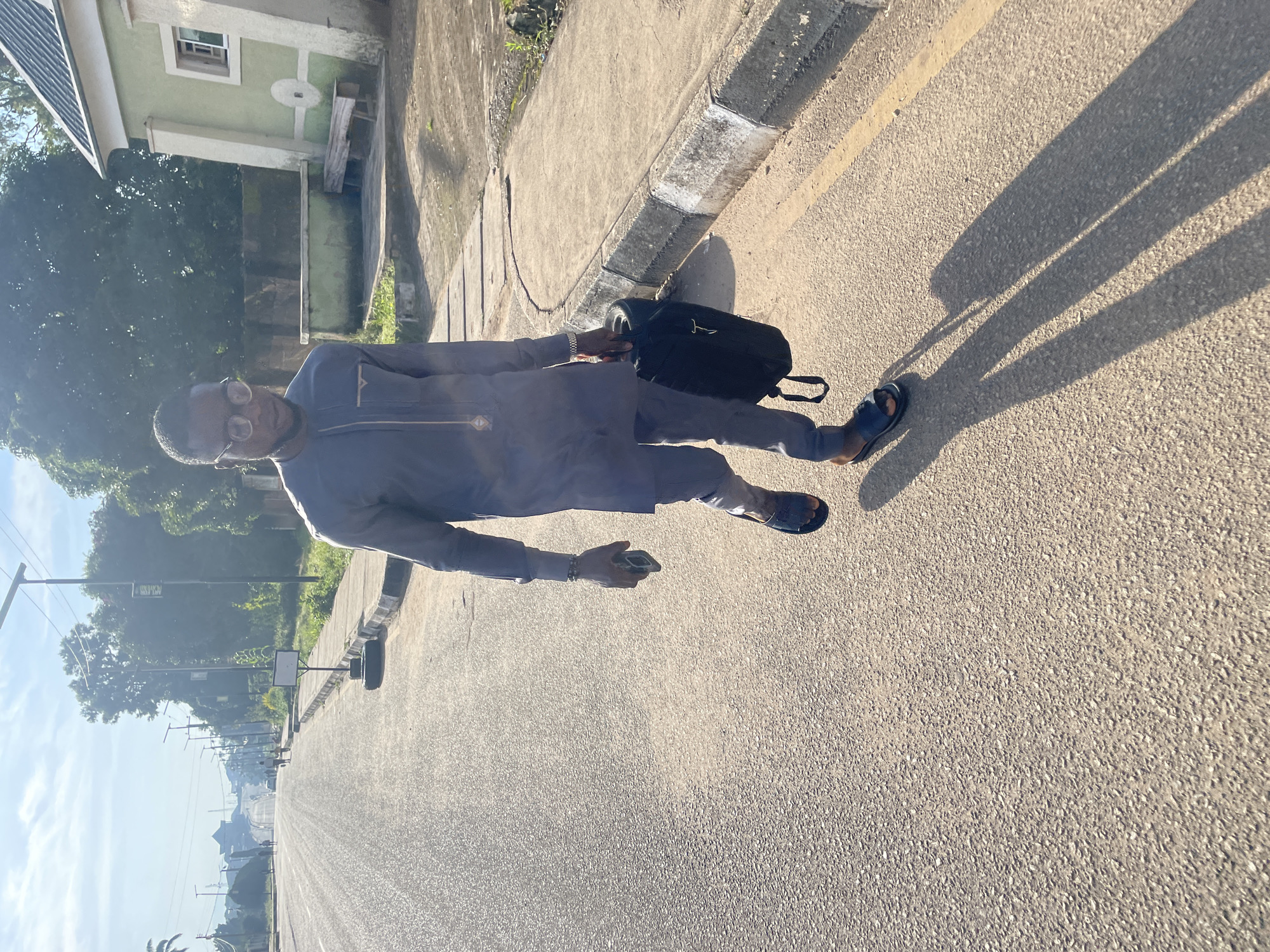 A man walks down a paved road in Jos, Nigeria, holding a black bag in his right hand and a cell phone in his left. He wears a grey tunic and trousers, and dark sandals. The sun casts long shadows across the road, indicating it is either early morning or late afternoon. To the right of the road, a light green building with a corrugated roof is visible, along with a wooden bench. Trees and other buildings line the street in the background, with streetlights and power lines overhead. The road appears to be a main thoroughfare, with other people and vehicles visible in the distance.