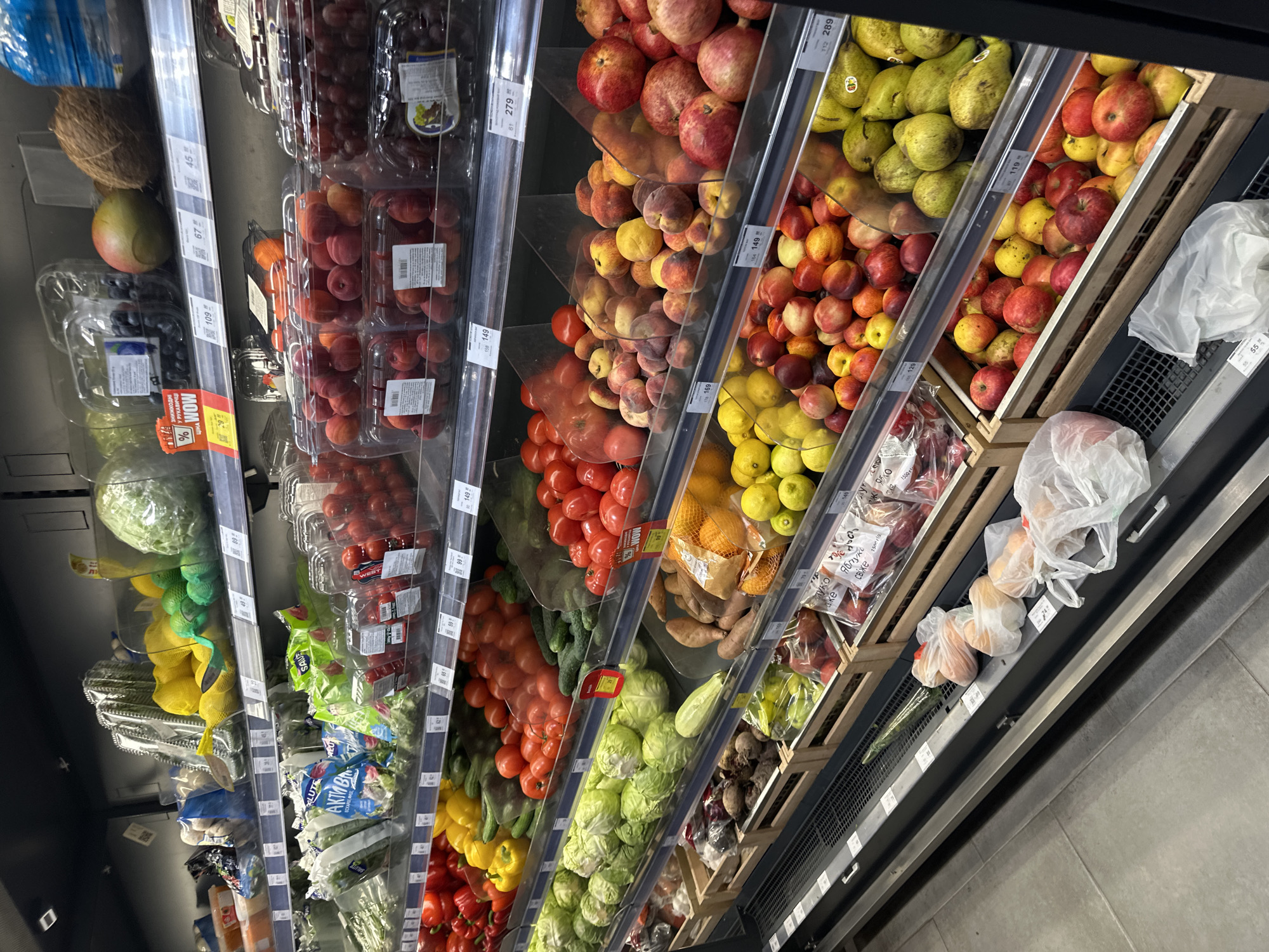 The image shows a grocery store produce aisle in Ternopil, Ukraine, with shelves stocked with various fruits and vegetables. On the top shelf, there are cabbages, lemons, avocados, and a coconut. The second shelf displays packaged produce like cucumbers, tomatoes, and bell peppers, along with loose grapes and apricots. The third shelf features more tomatoes, peaches, and apples. The bottom shelves hold cabbages, cucumbers, sweet potatoes, oranges, lemons, apples, and pears. Some items are in plastic bags or wooden crates. Price tags are visible throughout the display. The overall environment suggests a well-organized supermarket produce section.