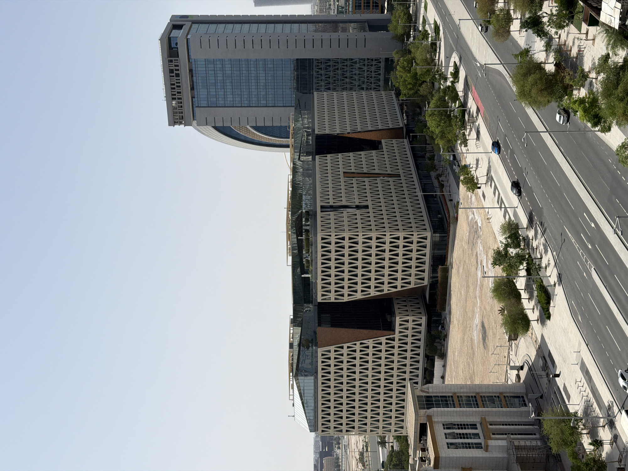 The image shows a high-angle view of a modern urban scene in Lusail, Qatar. Prominently featured is a contemporary building with a geometric latticework facade. A taller building with glass windows stands adjacent. A wide boulevard with multiple lanes runs alongside the buildings, with cars traveling on the road. Trees line the sidewalk between the road and the buildings. The sky is clear and pale blue.