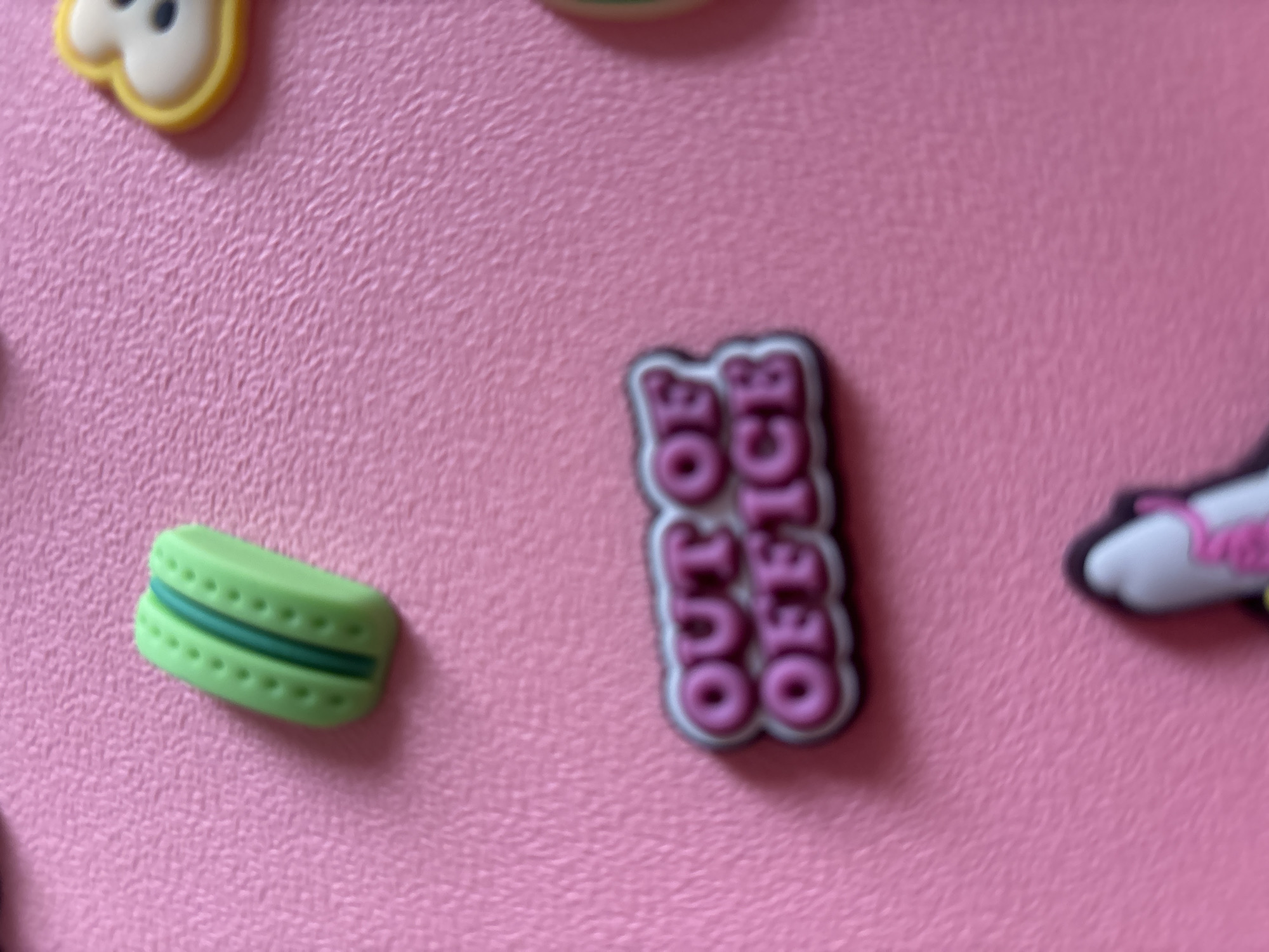 The image is a close-up shot of various charms scattered on a pink, textured surface. The charms include a green macaron, a pink "OUT OF OFFICE" text charm, and a portion of a yellow and white charm that appears to be a cartoon character's face. There is also a glimpse of a black and white charm with pink accents at the bottom of the frame. The lighting is even, and the focus is sharp on the charms. The background is a consistent pink texture. No specific location in Georgiyevsk, Russia is depicted or suggested by the image content.