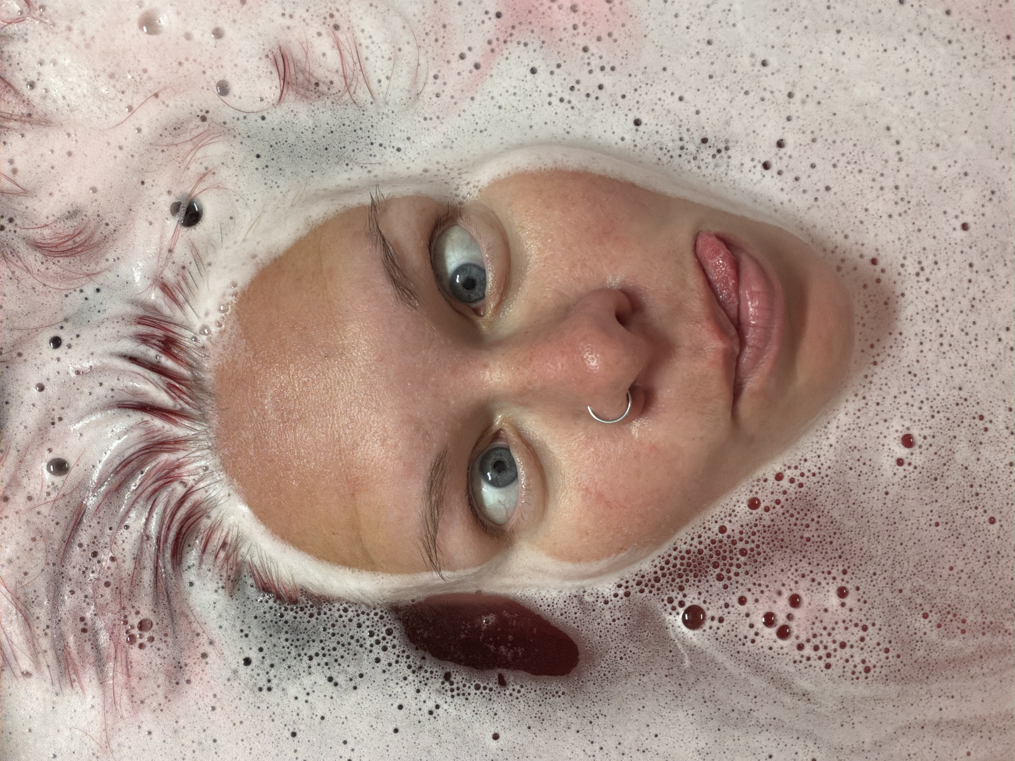 A close-up, slightly angled shot shows a person's face submerged in a bubbly, reddish-pink liquid. The person has fair skin, light blue eyes, and a silver nose ring. Their lips are slightly parted. Their hair appears to be dyed reddish-pink at the roots, fading into a lighter color where it meets the liquid. The liquid is foamy with many small bubbles. A darker, blood-red blotch is visible near the lower right side of the face. There is no information in the image to indicate that it was taken in the city of Chaikovsky, Russia.
