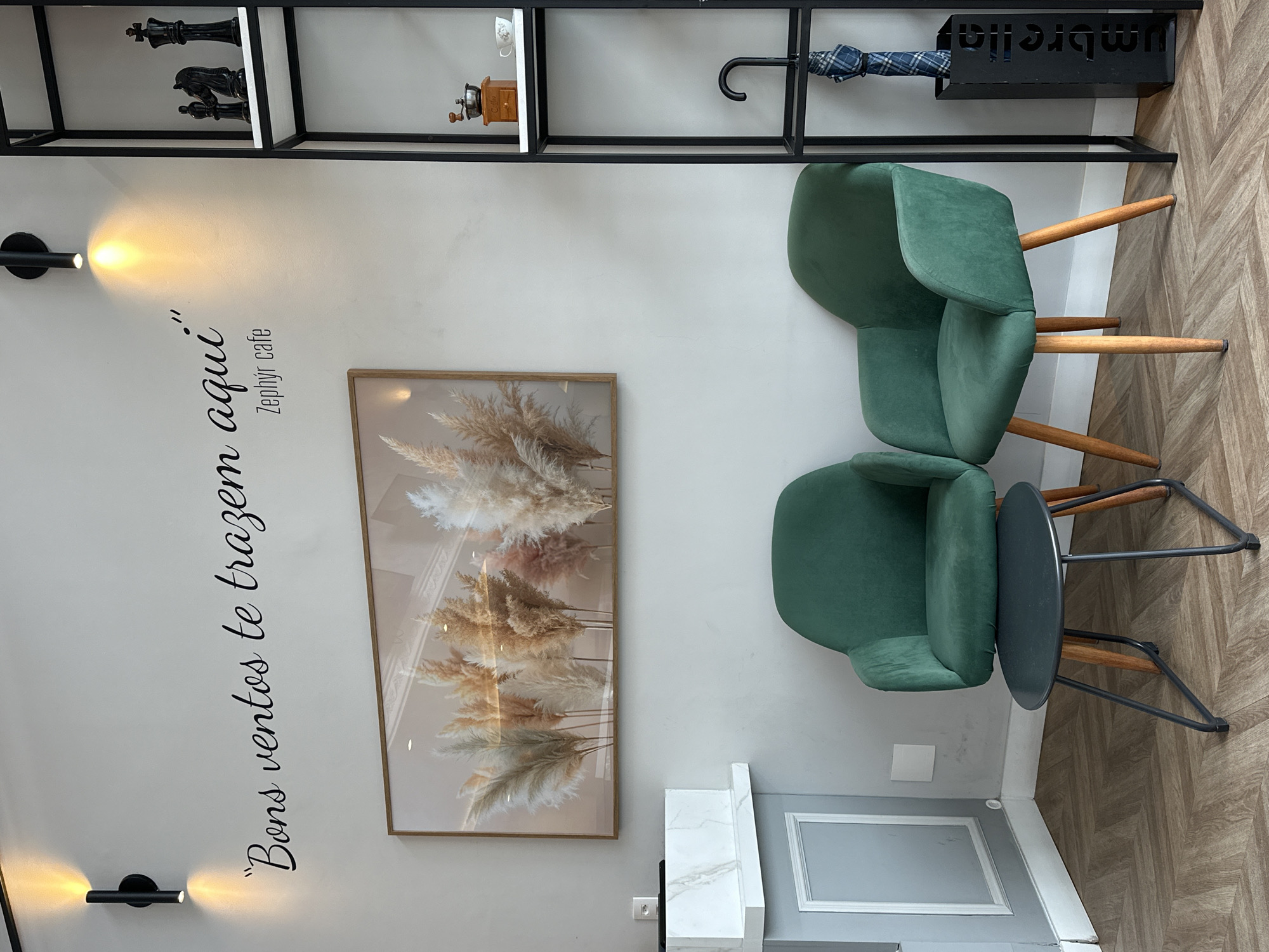 The image depicts the interior of a cafe in Florianópolis, Brazil, likely "Zephyr cafe" as indicated by the text on the wall. The scene is framed from a medium-wide shot, looking slightly upward, capturing a corner of the establishment. On the left, a pale gray wall features a black vinyl decal with the Portuguese phrase "Bons ventos te trazem aqui" (Good winds bring you here), followed by "Zephyr cafe". A modern, black, cylindrical light fixture is mounted above the text, casting warm light. Below the text, a framed piece of art displays a cluster of dried pampas grass plumes in soft, natural tones. To the left of the artwork, a white marble-topped console table with a gray base is visible, with a subtle electrical outlet on the wall behind it.
The focal point of the lower half of the image consists of two plush, emerald green armchairs facing slightly inward. Between them sits a small, dark gray, oval-shaped coffee table with a metal base and wooden legs. To the right of the chairs, a tall, black, minimalist shelving unit is positioned against the wall. The shelves hold a few decorative items: on the top shelf, a black chess set with pieces arranged, and a miniature wooden coffee grinder with a silver top, next to a small white teacup. The second shelf from the top has a white teacup. On the lowest visible shelf of the shelving unit, a black umbrella stand holds a folded umbrella with a blue and white checkered pattern. The floor is covered with a wood-look laminate flooring laid in a herringbone pattern. The overall ambiance suggests a modern, cozy, and stylish cafe setting.