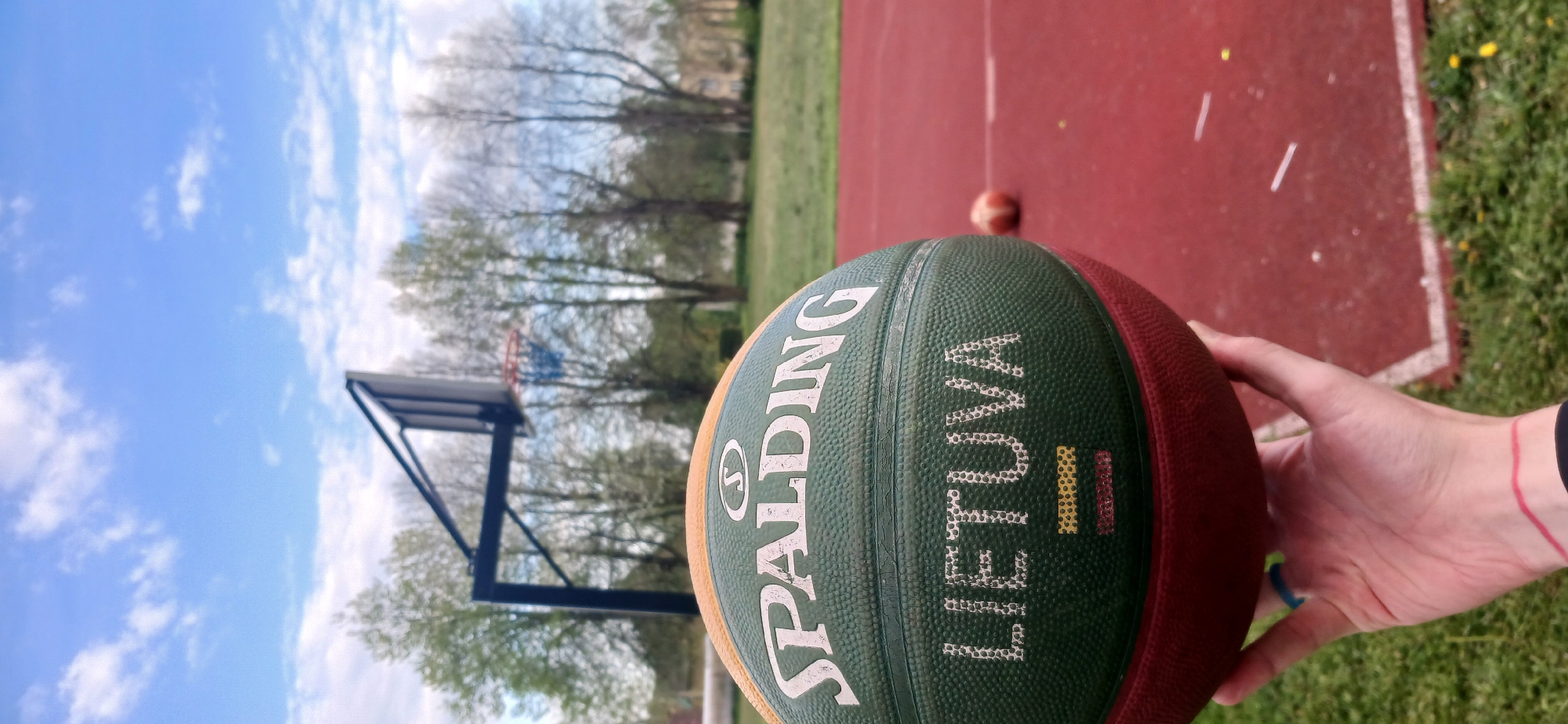 The image shows a Spalding basketball with the word "LIETUVA" (Lithuania) and a depiction of the Lithuanian flag. The basketball is held by a hand with a red wristband. Behind the basketball, there is a red basketball court with a basketball hoop, trees, and a cloudy sky. The location is likely Butkiškė, Lithuania, based on the inscription on the basketball.