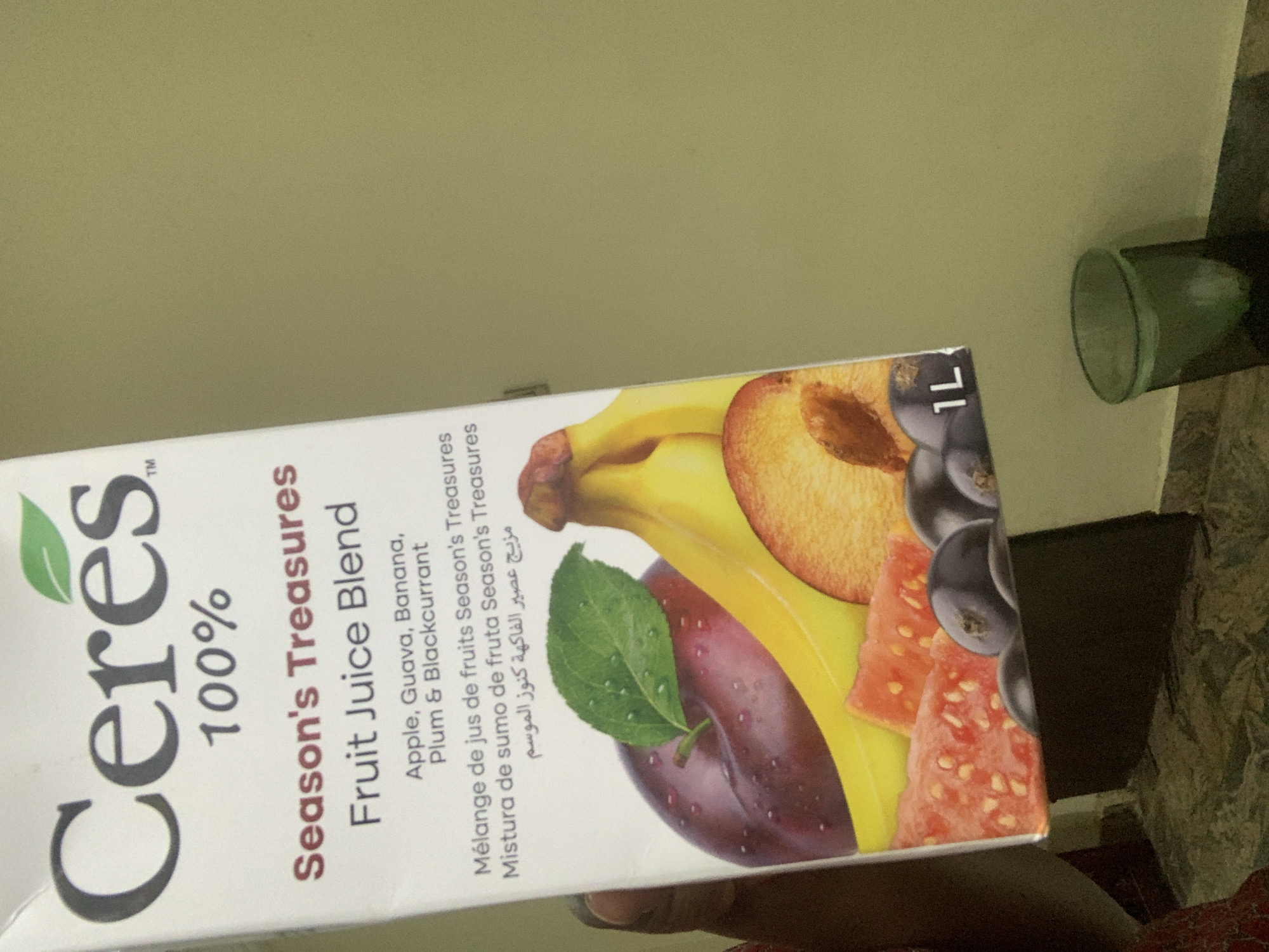 The image shows a carton of Ceres 100% Season's Treasures Fruit Juice Blend. The carton is white with the brand name "Ceres" in large, dark grey lettering at the top, accompanied by a green leaf logo. Below the brand name, "100%" is displayed in a smaller font. The text "Season's Treasures" is in dark red, followed by "Fruit Juice Blend" in dark grey. A list of fruits including "Apple, Guava, Banana, Plum & Blackcurrant" is also visible, along with translations in French, Spanish, and Arabic. The lower part of the carton features an illustration of various fruits, including a plum, bananas, a peach slice, guava chunks, and dark berries. In the bottom right corner of the carton, "1L" is printed.
The carton is positioned in what appears to be an indoor setting with a light olive-green wall in the background. In the foreground, there is a dark, patterned floor. To the right of the carton, a green, translucent glass is visible, containing some white material at the bottom. The lighting suggests an artificial light source, possibly from the side. The overall scene is consistent with a retail or domestic environment.
Although the image depicts a product commonly available in Abuja, Nigeria, there are no specific visual cues within the image that definitively place it within the city of Abuja. The image does not contain any people or specific geographical landmarks that would allow for precise location identification within Abuja.