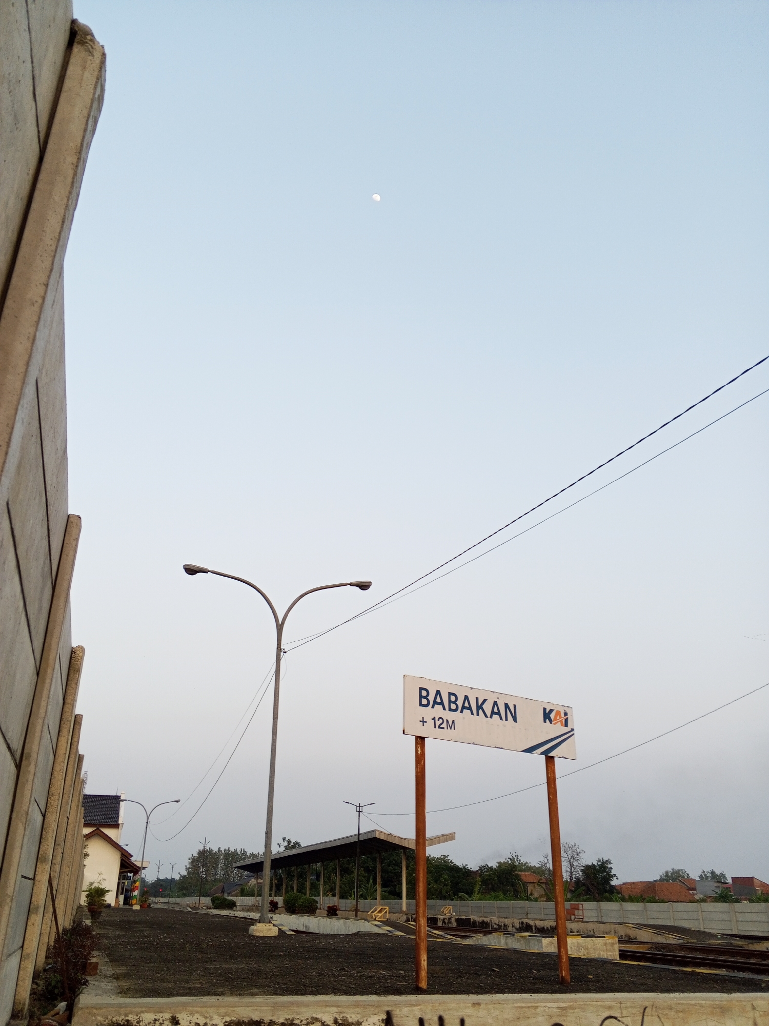 The image captures a serene outdoor scene, likely at a train station in the city of Pakusamben, Indonesia. The sky is a pale, uniform blue, with a faint, distant moon visible in the upper left quadrant. A large, concrete structure with a rough texture dominates the left side of the frame, casting a shadow on its surface. In the mid-ground, a double-headed streetlamp stands tall, its arms reaching out to illuminate the area.
Further into the scene, a white sign with blue trim reads "BABAKAN" and "+12M" alongside the "KAI" logo, indicating it's a railway station named Babakan, possibly at an elevation of 12 meters. The sign is supported by two rusty-orange poles. Behind the sign, a covered shelter with a dark roof and exposed wooden posts provides shade. The ground is covered with dark gravel and some construction materials, suggesting ongoing work or a simple, functional design for the station.
In the background, a cluster of houses with red-tiled roofs peeks through the trees, hinting at the surrounding residential area. A faint mist or haze is visible near the horizon, softening the distant landscape. The scene is devoid of people, creating a sense of quietude and stillness. The overall impression is that of an early morning or late afternoon setting, characterized by soft light and a tranquil atmosphere.