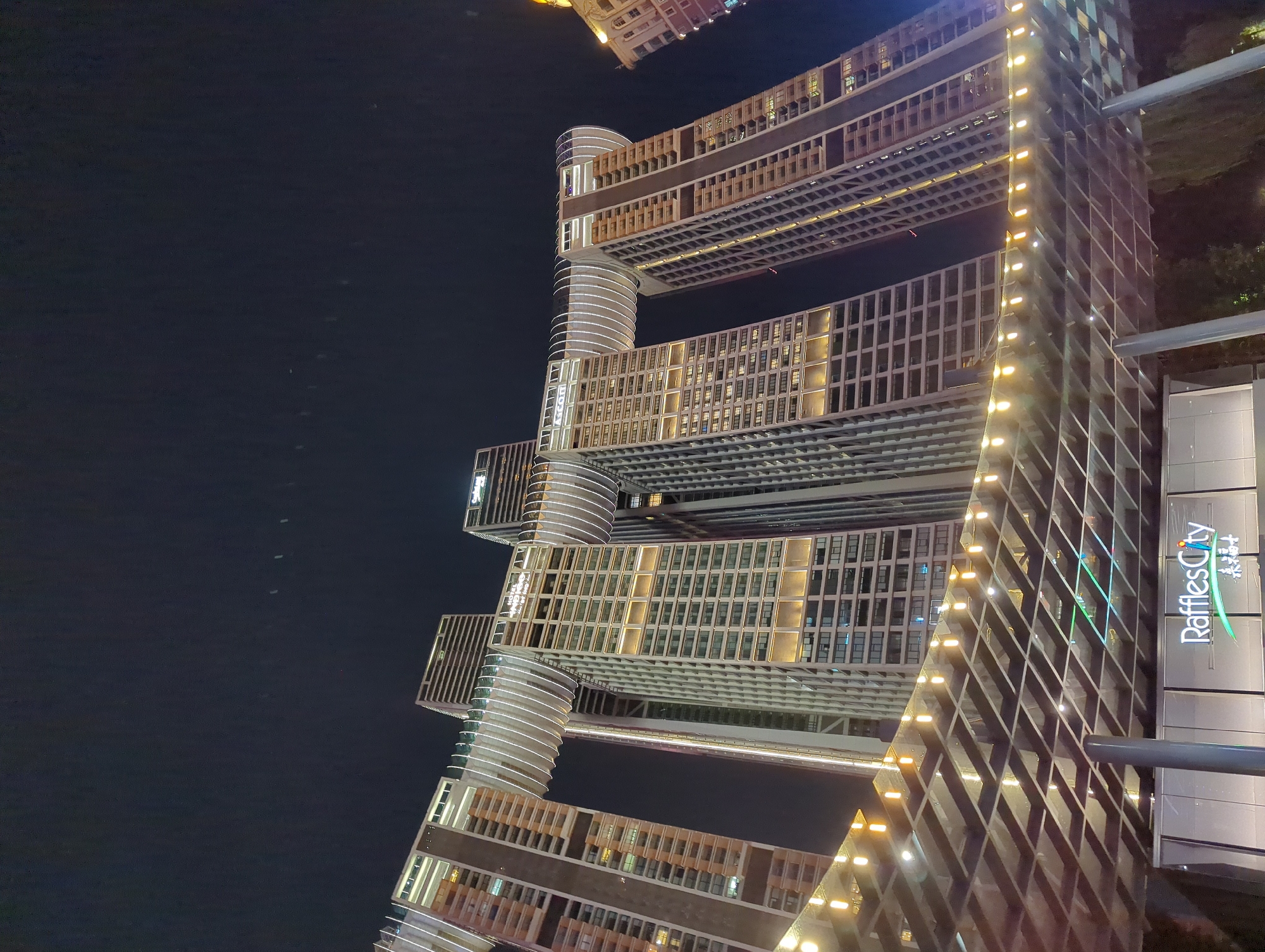 The image shows the Raffles City Chongqing complex at night, a distinctive architectural landmark in the Yuzhong District of Chongqing, China. The complex features multiple skyscrapers, some of which are connected by a horizontal structure resembling a ship or a dragon, illuminated by decorative lights. The sky above is dark, indicating nighttime. The foreground shows a covered walkway or entrance area with a geometric roof structure adorned with string lights. The "Raffles City" logo is visible on the building's facade, along with Chinese characters. The overall scene depicts a modern urban environment with impressive architecture and city lights.
