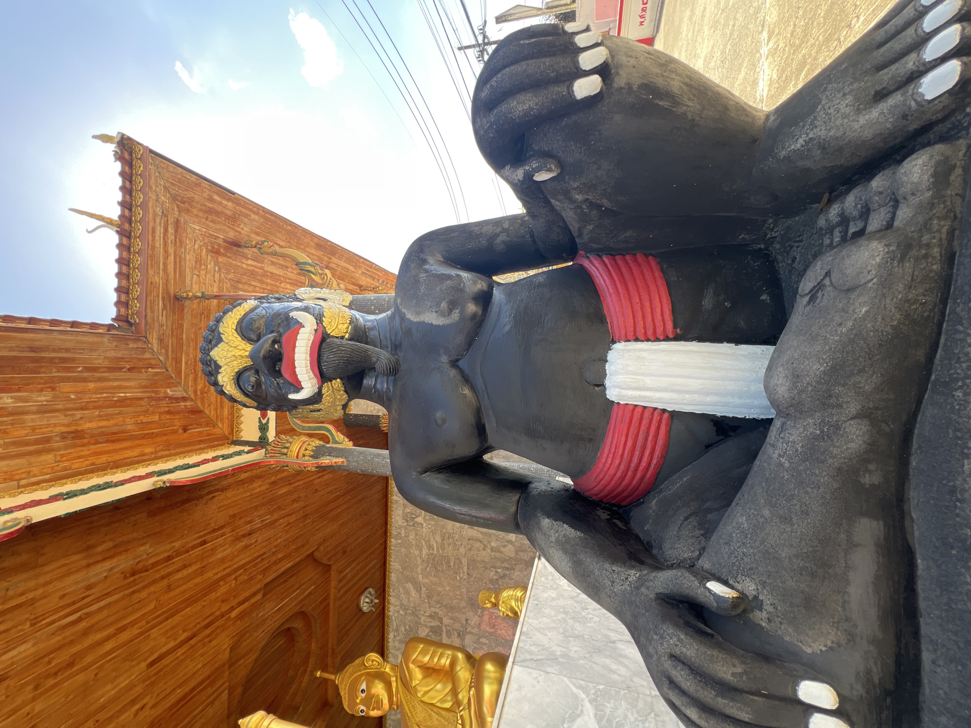 This media file shows a large, black statue of a fearsome figure with a wide, toothy grin, red lips, and gold embellishments around its face and neck. The figure is seated, with its knees bent and hands resting on its legs. It wears a red and white band around its waist. In the background, a wooden structure with decorative trim and a golden statue of Buddha can be seen. The sky is bright and sunny, suggesting it is daytime. The overall scene appears to be in an outdoor religious or cultural setting, possibly a temple complex, given the presence of the Buddha statue and the ornate architecture. The media file is oriented vertically, with the statue dominating the foreground. The location context provided is Thep Krasatti, Thailand.