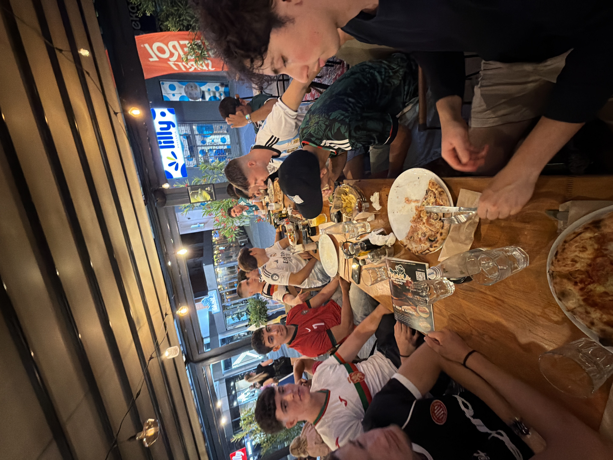 A group of young men are dining at an outdoor restaurant in Burgas, Bulgaria, seated around a wooden table laden with food and drinks. The restaurant appears to be covered by a dark awning, with string lights illuminating the area. Several of the men are wearing soccer jerseys, suggesting a possible connection to a sporting event or fan gathering. In the background, a "Lilly" drug store sign is visible, confirming the location in Burgas. The atmosphere seems casual and social, with the individuals engaged in conversation and enjoying their meal. The scene captures a moment of camaraderie and leisure in an urban setting.