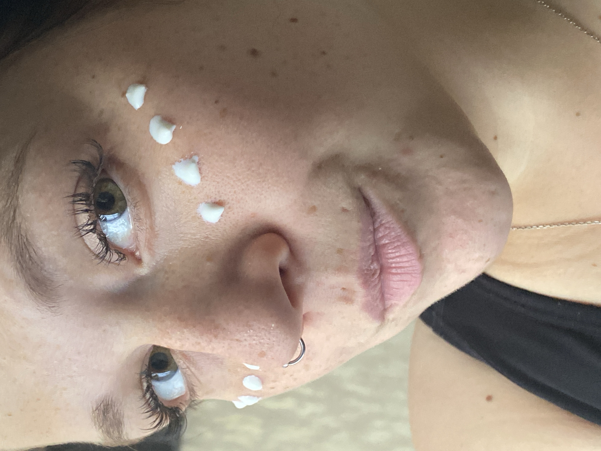 A close-up, medium shot shows a young woman with fair skin and freckles applying white cream to her face, with several dots of cream visible on her cheekbones. She has brown eyes, long dark eyelashes, and a nose ring. The woman is looking to her left and her lips are slightly parted. She is wearing a black tank top, and a silver necklace is visible. The background is out of focus and appears to be an indoor setting with a textured wall. The image does not contain any specific landmarks or details that would suggest a location in Chaikovsky, Russia.