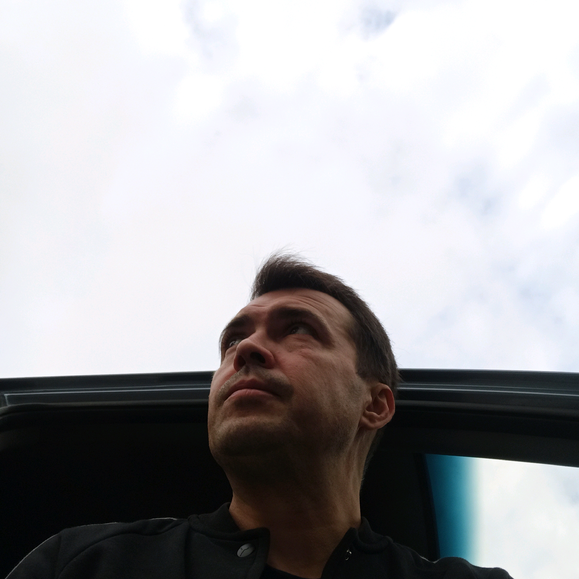 A man's face and upper body are visible in a close-up shot, looking upwards. He appears to be inside a vehicle, with the dark frame of a car roof or sunroof visible across the bottom of the frame. The sky, with some clouds, fills the background. The man has short, dark hair swept to the side, a light complexion, and is wearing a dark jacket. His expression is neutral or contemplative. The scene is likely captured outdoors in Nizhny Novgorod, Russia, given the provided context.