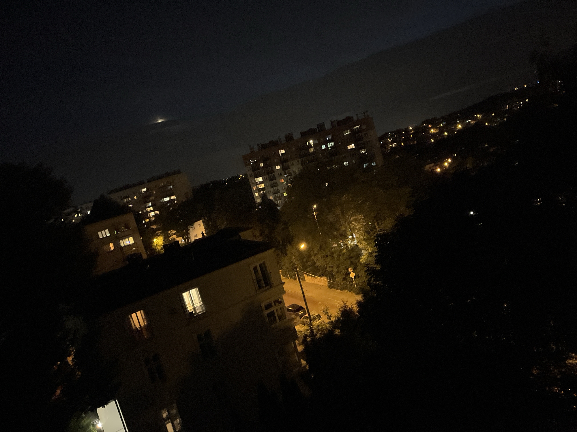 A nighttime aerial view captures a cityscape under a dark sky. Several apartment buildings are visible, some with illuminated windows, indicating occupancy. In the distance, a sprawling urban landscape is dotted with lights. A thin, faint light streak is present in the upper portion of the sky. The foreground is dominated by dark foliage, suggesting trees, which partially obscure the view of the buildings and the city. The image is angled, giving a diagonal perspective of the scene.