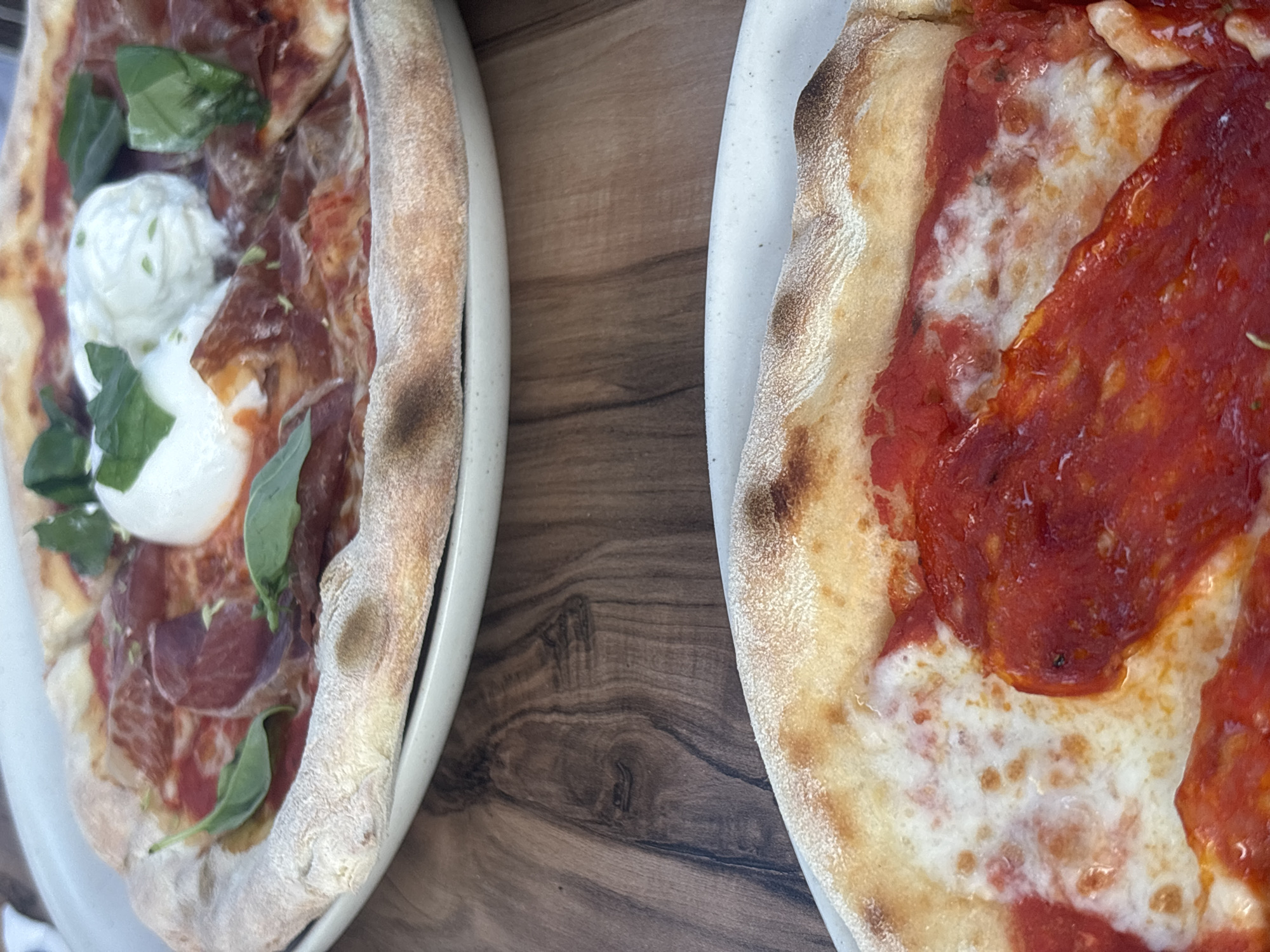 Two pizzas are presented on plates, with the top pizza being a prosciutto and burrata pizza, and the bottom pizza appearing to be a pepperoni pizza. Both pizzas have golden-brown crusts. The pizzas are placed on a wooden table. The overall scene suggests a casual dining setting, possibly a restaurant, in Castro Marim, Portugal.