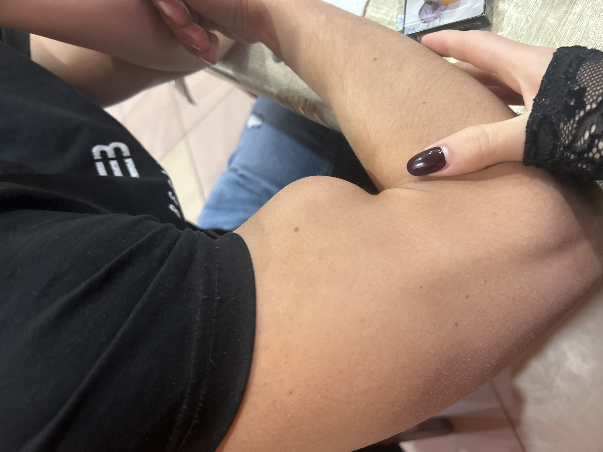 In Mirkovo, Bulgaria, an indoor scene captures an intimate moment involving two individuals. The primary focus is on the arm of one person, adorned in a black short-sleeved t-shirt, featuring a visible, flexed bicep. This arm exhibits light to medium skin tone with fine hairs and a few small moles or freckles. The second person's hand, covered by a black lace fingerless glove and featuring dark, possibly burgundy, polished fingernails, gently rests on the bicep of the first person.
The person wearing the t-shirt is seated, or at least positioned such that their torso, clad in the black fabric, is visible. A white, stylized logo on the t-shirt prominently displays what appears to be an uppercase "M" followed by two vertical bars (resembling "MII" or "MП"). In the background, hints of blue jeans with a torn detail suggest the lower body attire of the second individual. Further back, a light-colored surface, possibly a tiled floor or table, and a partially obscured object resembling a box or package are visible. Text on this object includes "SOBIYA" or "SOBIA". The overall setting appears casual and relaxed.
