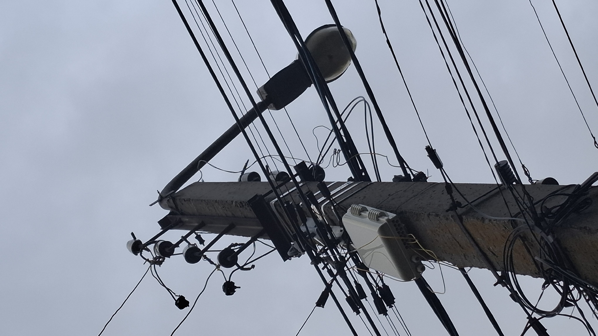 This image, taken in Rassvet, Russia, depicts a concrete utility pole set against an overcast, light grey sky. The pole is heavily laden with numerous overhead utility lines of varying thicknesses, predominantly dark in color. Towards the top, multiple white and dark ceramic or porcelain insulators are visible, securing several sets of electrical wires. Below these, a metallic bracket supports additional lines and components.
Mid-pole, a prominent white, rectangular enclosure, likely a telecommunications or fiber optic distribution box, is securely mounted. Several thin yellow wires emerge from the bottom of this box, intertwining with other dark cables that run vertically and horizontally along the pole. Further down, numerous black plastic strain relief clamps and cable bundles are attached, managing the intricate web of wires.
Extending horizontally from the right side of the pole is a dark-colored street light fixture, equipped with a large, oval-shaped lamp, illuminating the unseen area below. The pole itself appears weathered, exhibiting a rough, textured surface. No people, animals, or other distinct environmental features are visible in the frame, which focuses solely on the utility infrastructure.
