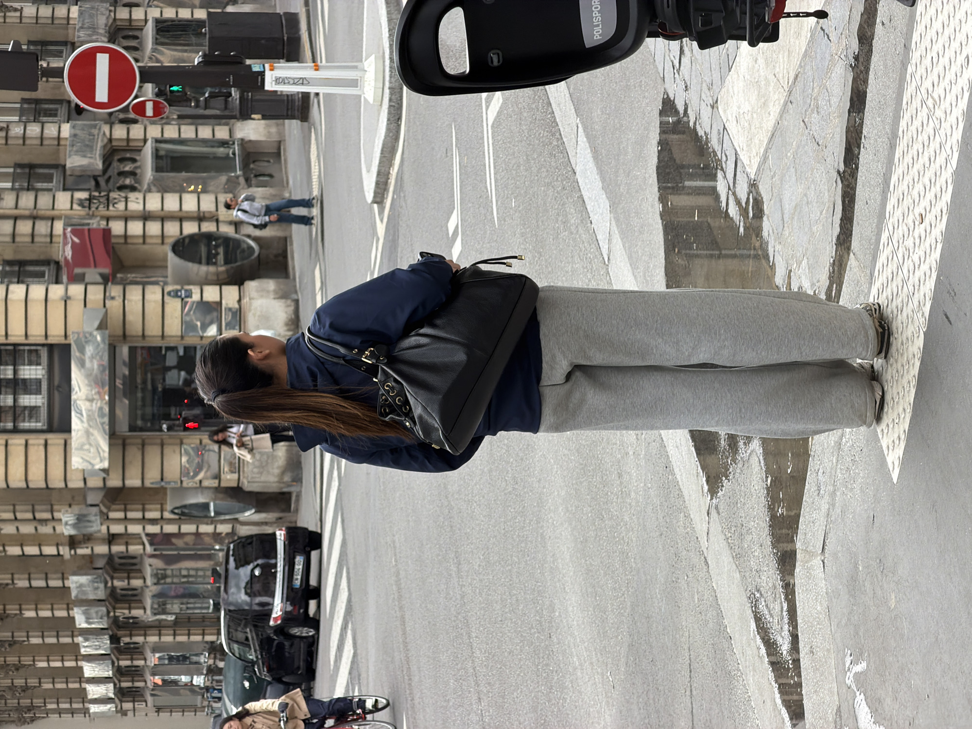 The image captures a street scene in Paris, France. A woman dressed in a navy jacket and grey sweatpants is walking along the street, carrying a black leather handbag. The street is paved and marked with white lines. A motorcycle is parked in the foreground. In the background, there are several cars and people, including a person on a bicycle. The buildings lining the street are typical Parisian architecture, with ornate facades and multiple windows. Street signs and traffic lights are also visible.