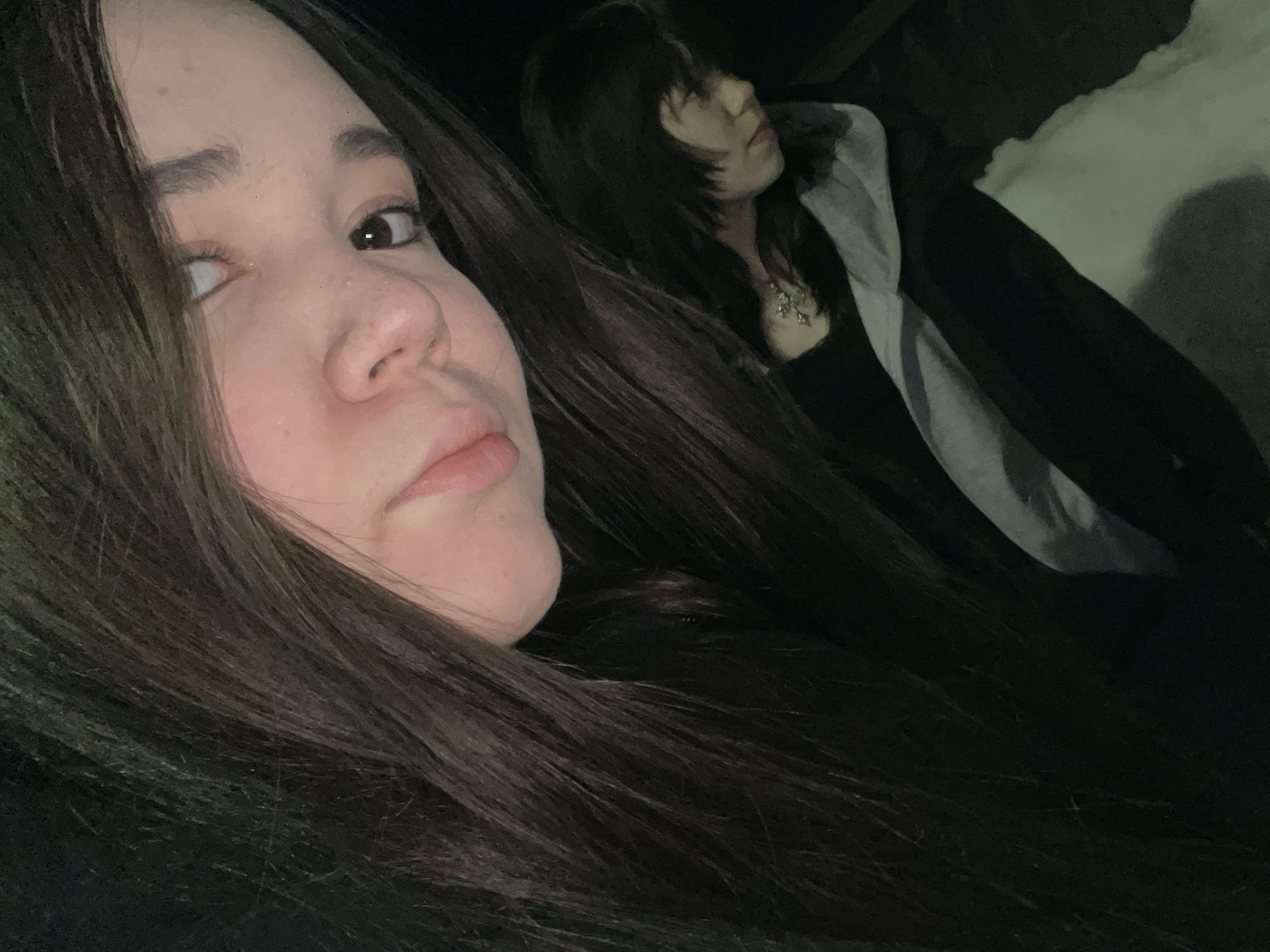 The image depicts two individuals within a dimly lit, undefined environment. The specific location within the city of Chaikovsky, Russia, is not discernible from the visual information provided.
In the foreground, an individual's face and upper chest occupy the upper left portion of the frame, angled towards the viewer. This person has long, straight, dark brown hair that cascades down the left side. Their face exhibits a neutral expression with slightly parted lips and dark eyes looking towards the right. Direct lighting illuminates their face, suggesting a close-range light source, possibly a camera flash.
In the middle ground, positioned to the lower right and slightly out of focus, is a second individual. This person has shorter, dark hair, styled with layers or bangs, and is observed from a side profile, looking downwards and to the right. They are wearing a dark top, and a metallic necklace with a prominent, complex pendant (possibly a cross or star-shaped design) is visible on their chest. A light-colored, possibly white or light grey, fabric item is draped over their lap or lower torso.
The background is uniformly dark and lacks any discernible features, indicating low light conditions or a shallow depth of field. No specific actions or interactions beyond their postures are evident.