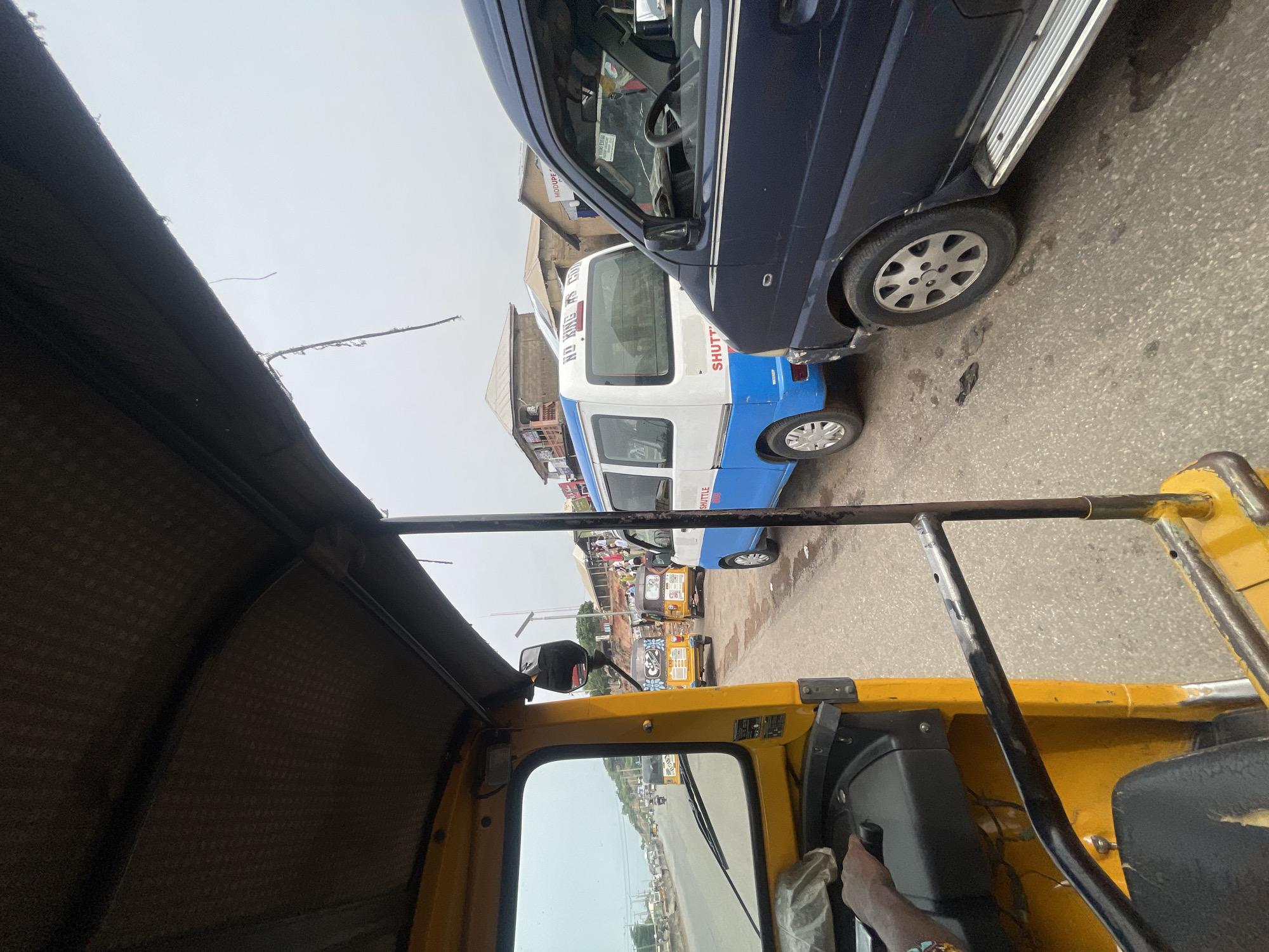 The image, likely taken in Lokoja, Nigeria, shows a view from inside a yellow auto rickshaw, also known as a "keke". In front, there's a blue van parked on the right side of the road. A white and blue shuttle bus is also visible beyond the van. The street appears to be busy with other kekes and people. The sky is overcast. The photo captures a typical urban scene of public transportation and traffic.