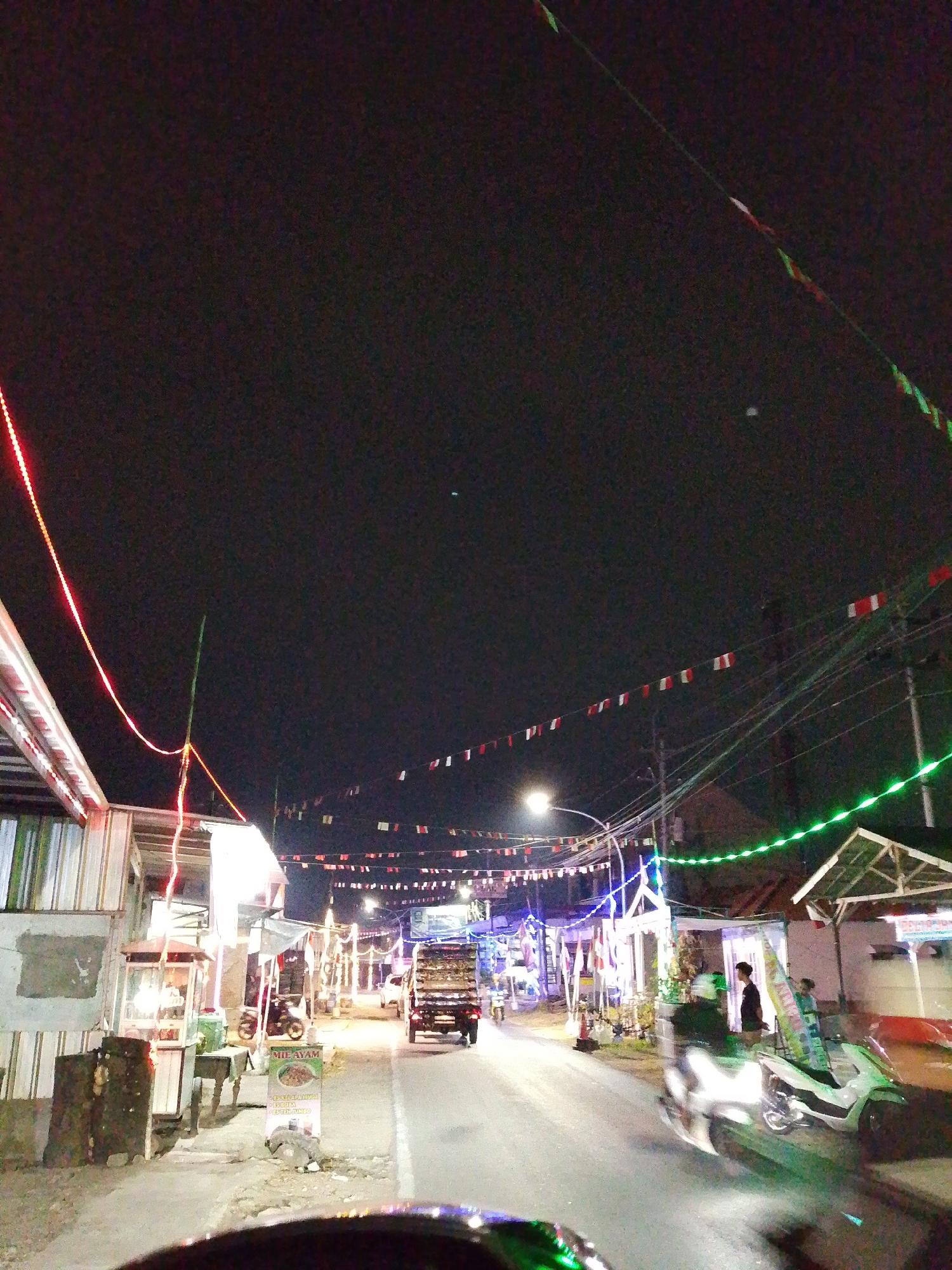 The image depicts a street scene at night in Pagongan, Indonesia, characterized by festive decorations and illumination. Red and white triangular flags are strung across the street, along with green and red string lights. Buildings line both sides of the road, many adorned with bright lights.

On the left side of the street, there are several structures, including what appears to be a food stall with a glass display and seating outside. A sign for "MIE AYAM" is visible, indicating a noodle dish. A truck with a flatbed loaded with goods is driving down the road from the distance.

On the right side of the street, there are more buildings and a white motorcycle parked. A person is standing near the motorcycle, and another person is visible inside a doorway. The street is paved and appears to be a main thoroughfare. The overall atmosphere suggests a lively and possibly celebratory evening in the town.