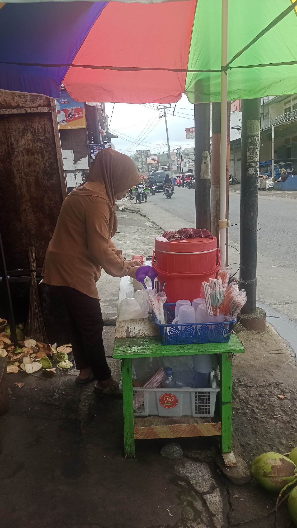 A woman wearing a light brown hijab and blouse stands behind a small green wooden table displaying drinks and supplies. She is preparing a beverage, holding a purple pitcher over a plastic cup. To her right, a large red insulated cooler sits on the table, topped with a red cloth. In front of the cooler, a blue plastic basket holds several white plastic cups and numerous individually wrapped straws. Below the table, a white plastic crate contains more straws and a water bottle, with a red logo on the front of the crate. To the left of the table, a pile of cut coconuts and husks are scattered on the ground. A broom leans against a weathered, dark-colored structure to the far left. The scene is outdoors, with a paved street visible behind the table, featuring several motorcycles and cars. Tall poles with electrical wires are present, and buildings line the street in the background. A large, colorful umbrella, sectioned into purple, red, and green, is held up by poles, providing shade over the area. The ground appears wet and somewhat dirty. This street scene is located in Karyamandala, Indonesia.