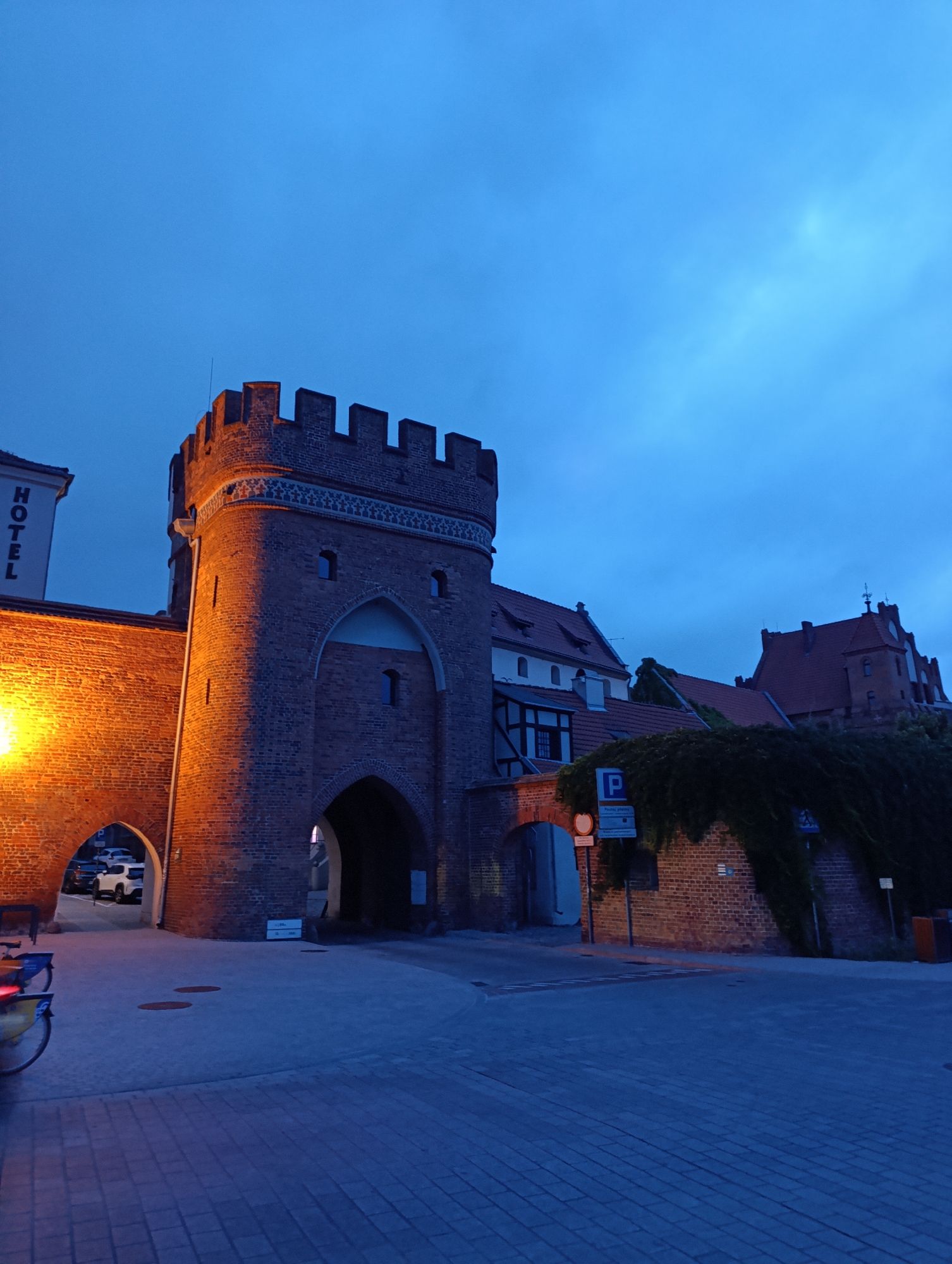 The image depicts the historic Gate of Scotland (Brama Szkocka) in Toruń, Poland, at dusk. The medieval brick gatehouse, illuminated by warm artificial light, stands prominently on the left side of the frame. A large archway leads through the gate. To the left of the gatehouse, a building with the word "HOTEL" vertically displayed on its facade is visible. In the background, more brick buildings with red-tiled roofs, characteristic of Toruń's Old Town, are visible, some covered in ivy. A street with cobblestone paving stretches in front of the gate. A white car is parked under an adjacent archway. A bicycle is parked on the left side of the foreground. The sky is a deep blue, indicating twilight. There are no people actively engaged in any actions.