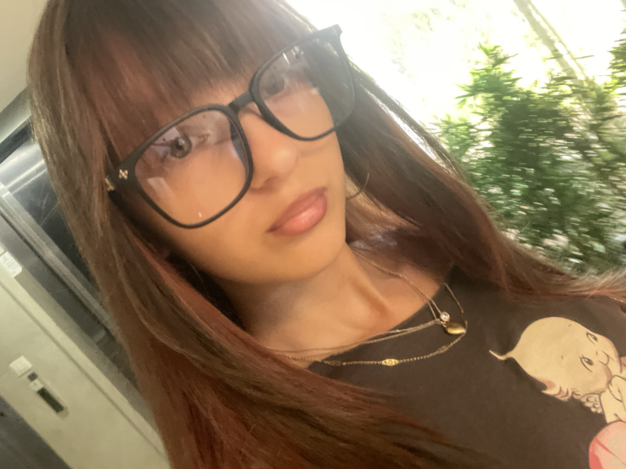A medium shot captures a young woman with long, reddish-brown hair, wearing glasses and a dark t-shirt with a baby graphic. She is indoors, as indicated by the metallic and white horizontal surface visible in the upper left quadrant of the image, possibly a kitchen counter or cabinet. The background on the right features a bright, out-of-focus view of lush green foliage, suggesting proximity to a window or outdoor space. The woman is angled towards the camera, with her face partially obscured by her hair and glasses. She is wearing two delicate necklaces. The lighting suggests it is daytime. The image does not provide any specific visual cues to place it within the city of Raanana, Israel.