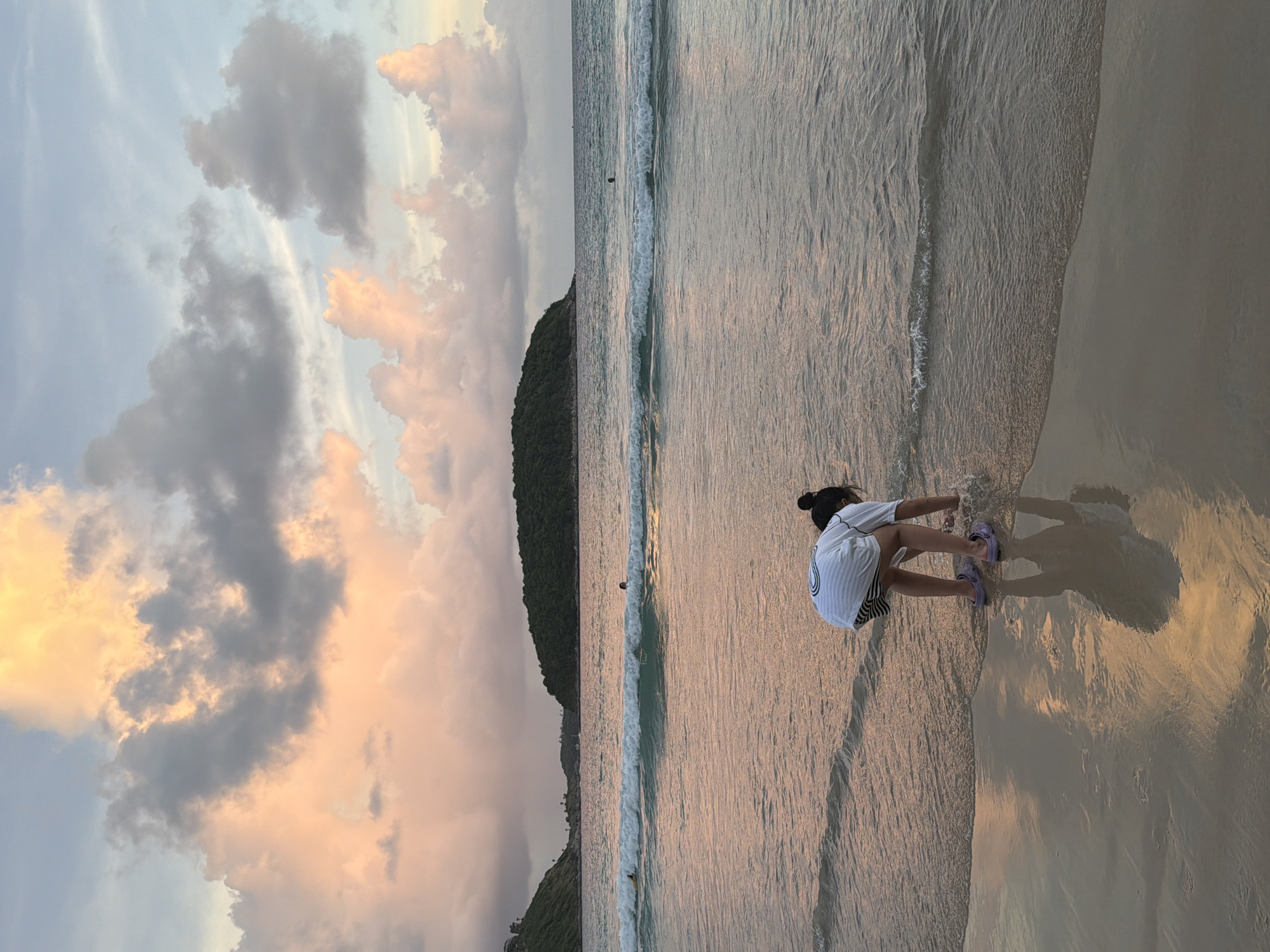 In Rawai, Thailand, a young person wearing a white t-shirt and striped shorts is crouched on a sandy beach, reaching into the shallow water. Their reflection is visible on the wet sand. The ocean has gentle waves, and in the distance, a lush green island juts out into the sea. The sky is filled with dramatic clouds, tinged with shades of pink and orange from the setting sun. Other people can be seen swimming or wading in the water further out.