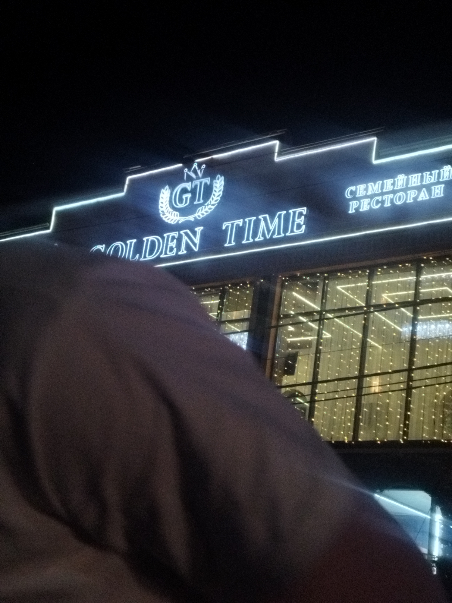 The image shows a neon sign at night that reads "GOLDEN TIME" in English and "СЕМЕЙНЫЙ РЕСТОРАН" in Russian, which translates to "FAMILY RESTAURANT". The sign is lit with white neon lights against a dark background. Below the sign, a building with large windows is visible, decorated with strings of small, white lights. A dark, out-of-focus object, possibly clothing or fabric, occupies the foreground, obscuring a portion of the building and the sign. The location is identified as Shymkent, Kazakhstan.
