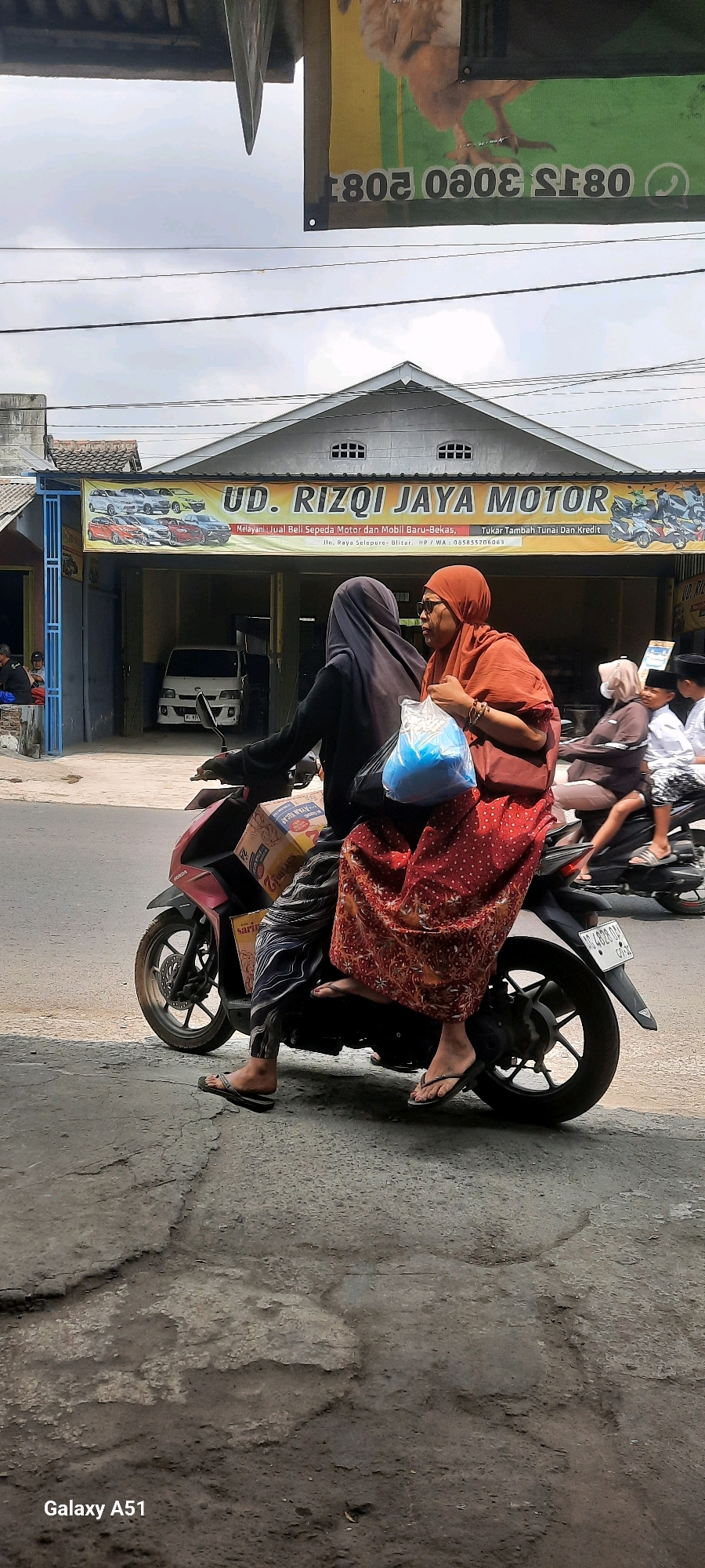 In Selopuro, Indonesia, two women are riding on a red and black scooter. The woman in the front, wearing a black hijab and patterned pants, is driving. The woman behind her, wearing an orange hijab and a red patterned skirt, holds a blue bag in her left hand. They are riding past a building with a yellow banner that reads "UD. RIZQI JAYA MOTOR" and displays images of cars and motorcycles. A white truck is visible inside the building's garage. In the background, another motorcycle carries a woman and a child. The road surface appears rough and uneven, with some patches of visible concrete. The sky is overcast.