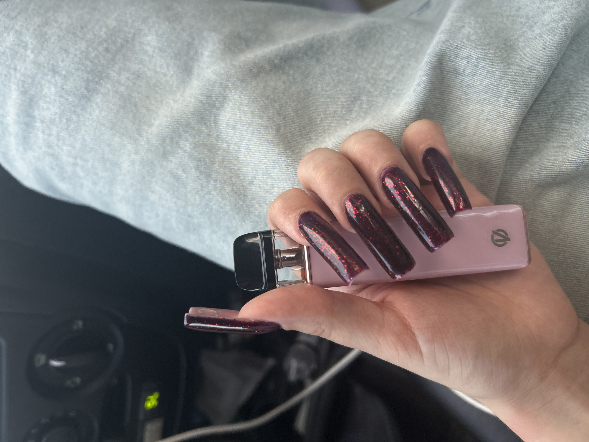 A person's hand with long, dark red glitter nails holds a light pink vape pen. The person is wearing light blue denim jeans. The background is dark and blurred, showing what appears to be the interior of a car with a black dashboard and circular knobs. The scene is likely indoors, possibly inside a vehicle, and there is no discernible connection to the city of Chaikovsky, Russia.