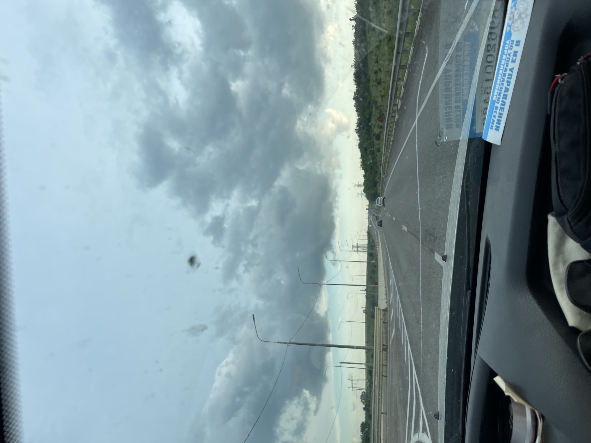 The image is a view from the driver's seat of a vehicle, looking through the windshield at a highway. The sky is overcast with dark clouds. The highway has multiple lanes and is bordered by guardrails and a grassy median with trees. There are streetlights visible along the side of the road. In the distance, there are power lines and electrical towers. A few other vehicles are on the highway. A blue and white sticker or document is affixed to the inside of the windshield, partially obscuring the view. The dashboard and part of the steering wheel are visible in the foreground.