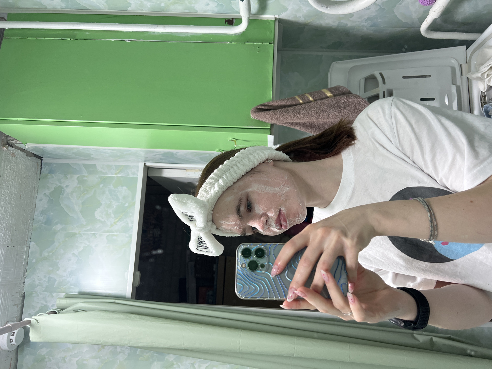 A person is in a bathroom in what appears to be Chaikovsky, Russia. The bathroom has green walls and a green cabinet above the mirror. The person is wearing a white t-shirt and a white headband with a bow, and has a face mask on. They are holding a smartphone and taking a selfie in the mirror. A brown towel is hanging nearby. The bathroom has a light green shower curtain and a white laundry basket. The background shows a typical, older bathroom interior.