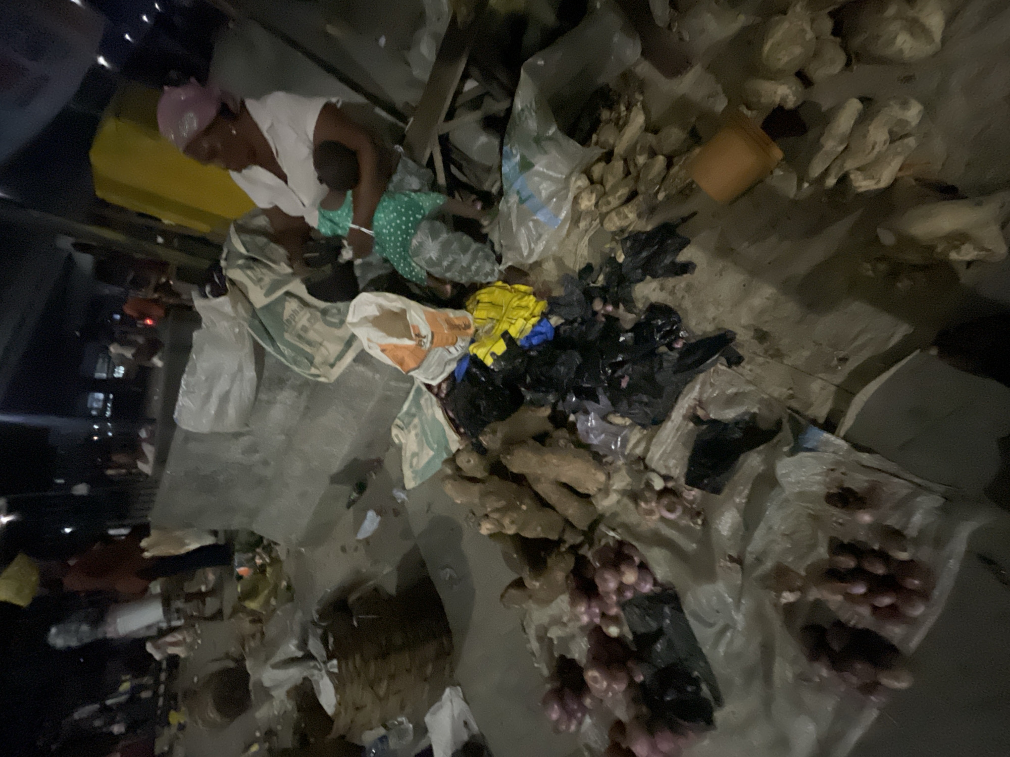 The image captures a dimly lit, informal market scene at night in Lekki, Nigeria, viewed from a high, tilted angle. The central subjects are an adult woman and a small child. The woman wears a light-colored top and a pink head covering, leaning forward as if attending to goods on the ground. A young child, dressed in a green and white polka-dotted outfit, is positioned close to her.
The ground, likely concrete or pavement, is covered with various produce and market items. Piles of reddish-purple onions or shallots are visible, some bundled in clear plastic bags. Numerous gnarled, light-brown roots, resembling ginger or other tubers, are also laid out. Other items are packed in black plastic bags.
Several large, utilitarian sacks are scattered around, some appearing to be repurposed. One prominent sack bears partially legible text, "ALPHA CEM," suggesting it was originally a cement bag. A small orange bucket and other colorful bags (yellow and orange) contribute to the cluttered market environment. The scene is illuminated by artificial light sources, confirming the nighttime setting and casting shadows. The activity centers around the woman working at her produce stall, with the child accompanying her. The bustling yet informal nature of the market is evident. Emotions are not clearly discernable due to the lighting and angle, but the woman appears focused on her task.