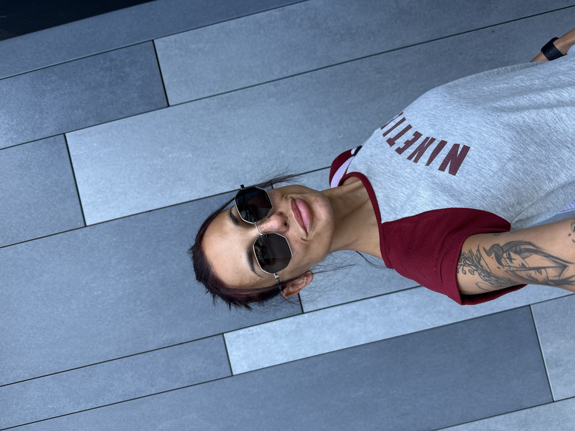 A woman is taking a selfie outdoors, standing in front of a textured gray wall. She is wearing large, hexagonal sunglasses, a gray t-shirt with burgundy sleeves, and a burgundy graphic on the front that reads "NINETI". She has dark hair pulled back and visible tattoos on her left arm. The wall appears to be made of rectangular panels of varying shades of gray and blue. The image does not contain any information that would allow for the identification of the city of Pleven, Bulgaria, or any other specific location.