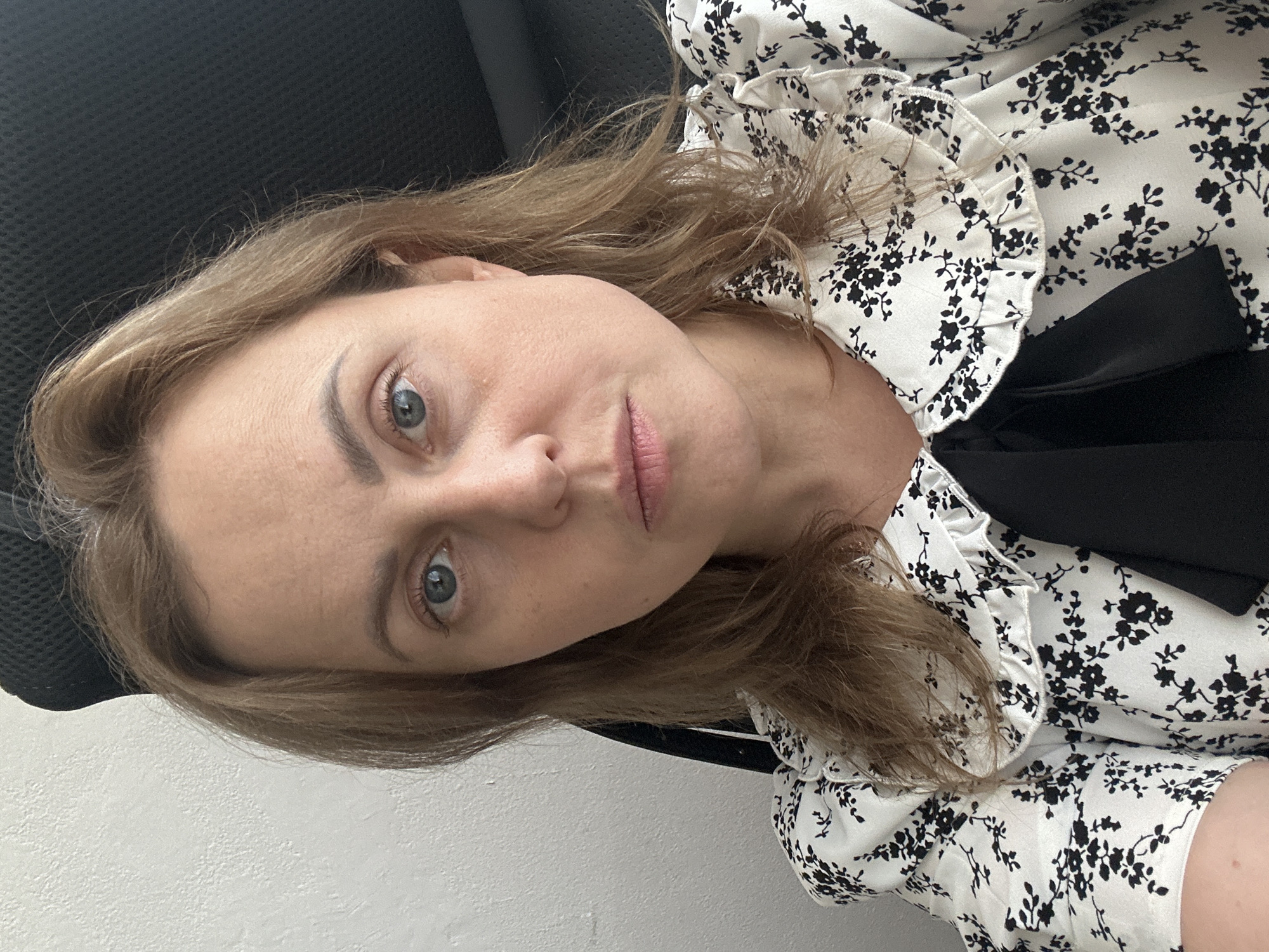 A woman with light brown hair and blue eyes is looking directly at the camera. She is wearing a white blouse with a black floral pattern and a black bow at the neck. The background is a textured white wall and a dark seat back. The image was taken indoors, likely in an office or home environment in Sochi, Russia.