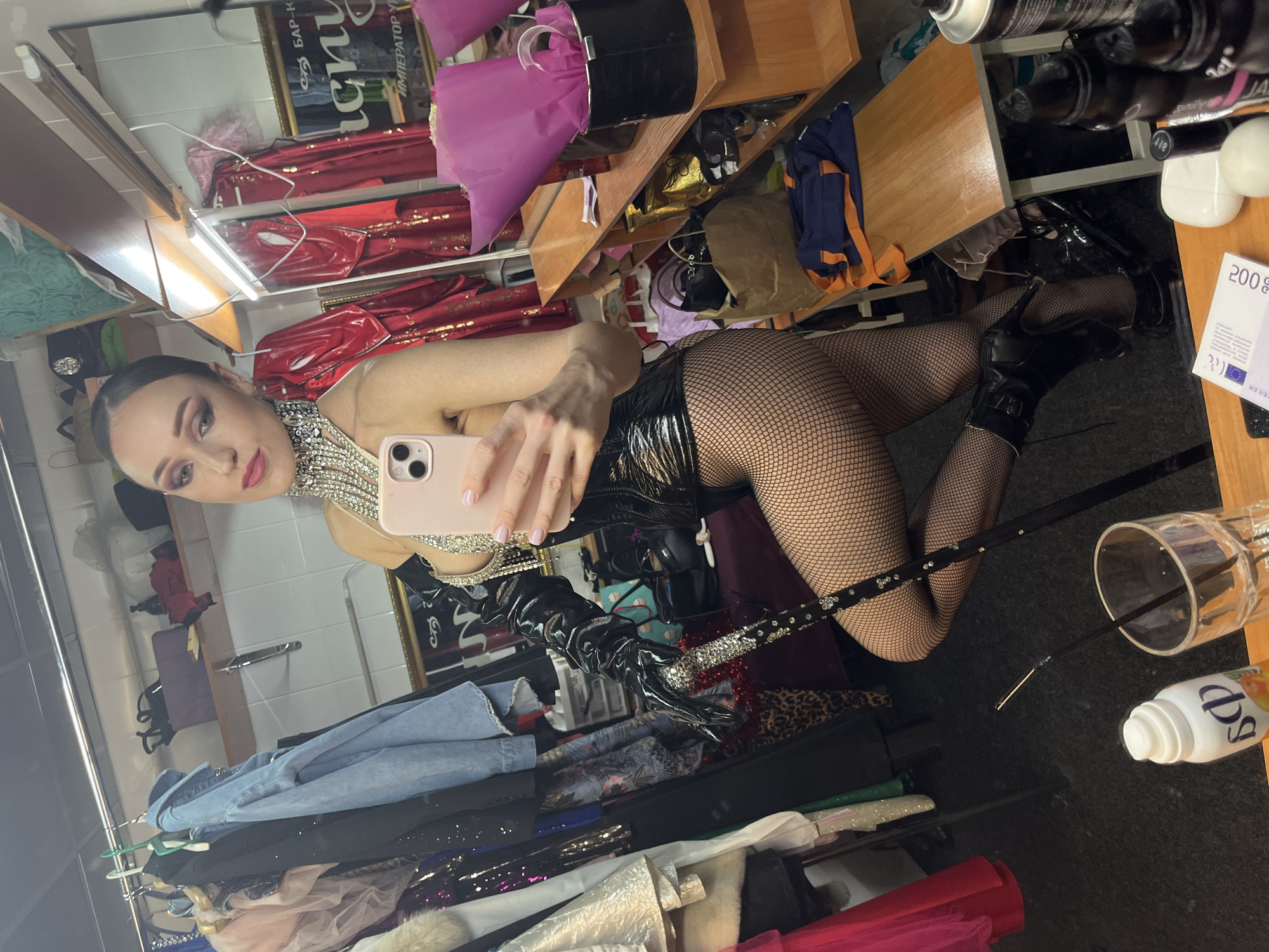 A woman is taking a selfie in a dressing room. She is wearing a black corset, black fishnet stockings, and black high-heeled boots. She is also wearing long black gloves and a sparkly necklace. She is holding a cane in her right hand and a pink smartphone in her left hand. The dressing room is cluttered with clothes hanging on racks and shelves filled with various items, including makeup, hair products, and a bouquet of flowers. A mirror on the wall reflects the scene. The background includes a framed poster with Cyrillic text that appears to be in Kazan, Russia, possibly indicating the location of the establishment.