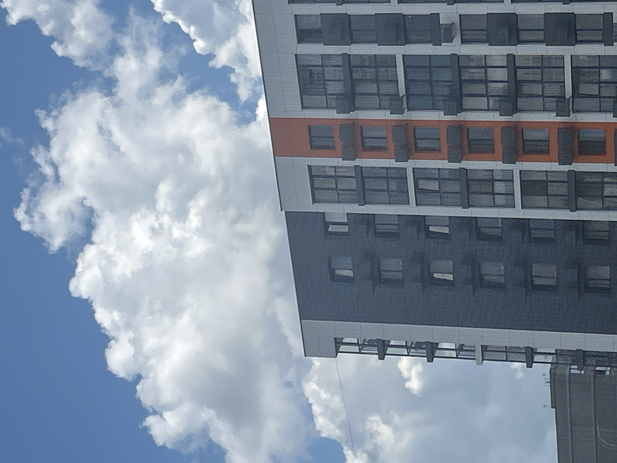 The media file depicts the exterior of a modern, multi-story building against a backdrop of a bright blue sky filled with fluffy white clouds. The building has a segmented facade, with sections of light grey paneling and dark grey cladding. A prominent horizontal band of orange accents runs across the middle section of the building, breaking up the color scheme. The windows are numerous and appear to be large, rectangular panes, some with dark frames. The building's design suggests a residential or office complex.
The composition is angled, with the building occupying the right side of the frame and the sky and clouds the left. The lighting suggests it is daytime, likely midday or afternoon, given the brightness of the sky and the clarity of the clouds. The weather appears to be fair and sunny. There are no people or specific activities visible, the focus being on the architecture and the natural elements. The image gives a sense of urban development in a clear, open sky setting. There is no visible text in the image. The location context provided is Orlovskii, Russia.