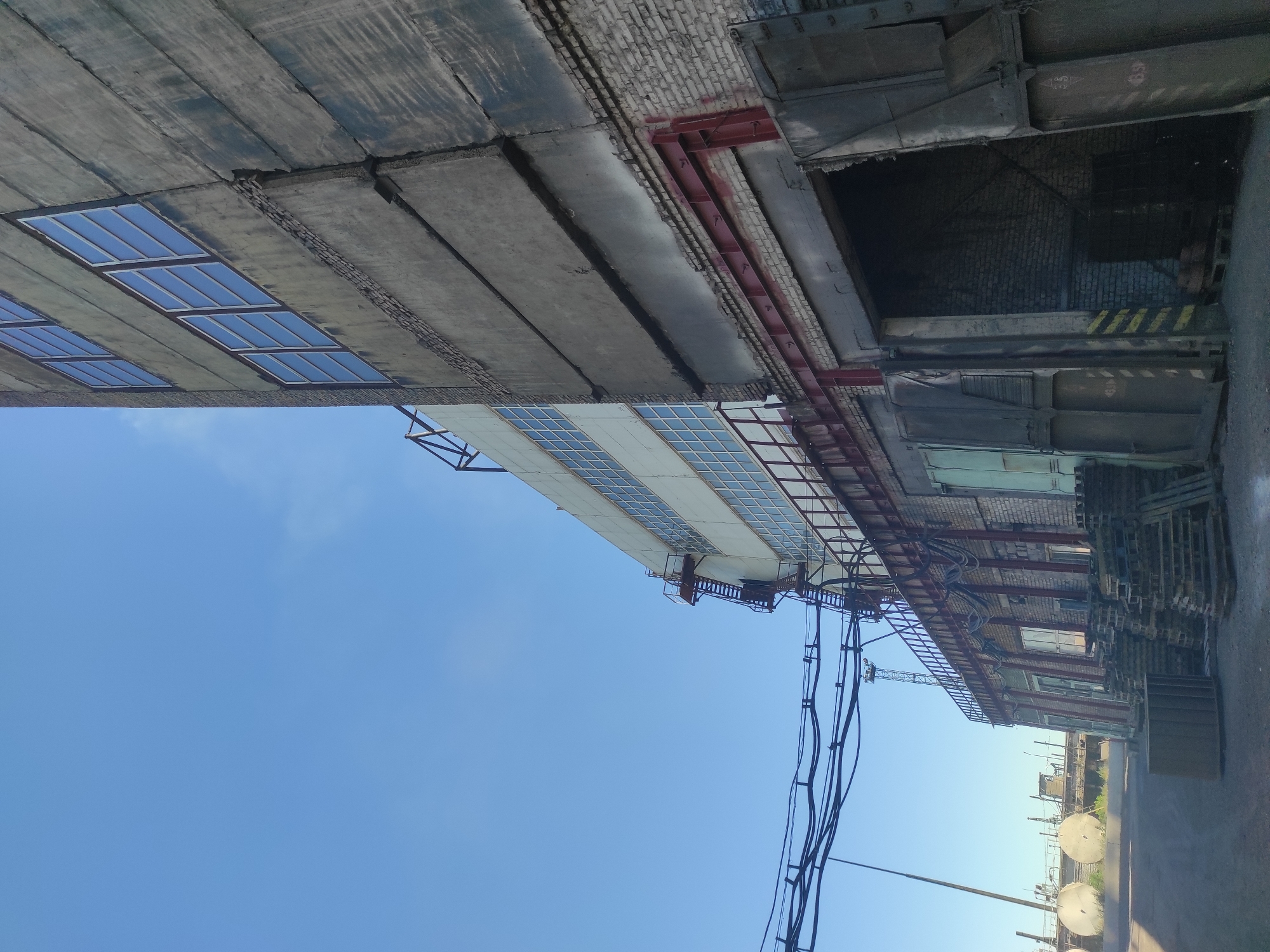 The image depicts an industrial building with a clear blue sky in the background. The building features a multi-story structure made of concrete and brick, with rows of windows on the upper levels. A metal fire escape is visible on one side of the building. In the foreground, there is an open loading bay with heavy industrial doors. Pallets of materials are stacked near the loading bay. Electrical cables are strung across the upper part of the scene. In the distance, industrial elements and what appear to be large circular structures are visible, along with a construction crane. The overall impression is one of an active industrial or manufacturing site. This scene is not in the city of Kyzyl, Kazakhstan; it is likely an industrial area elsewhere.