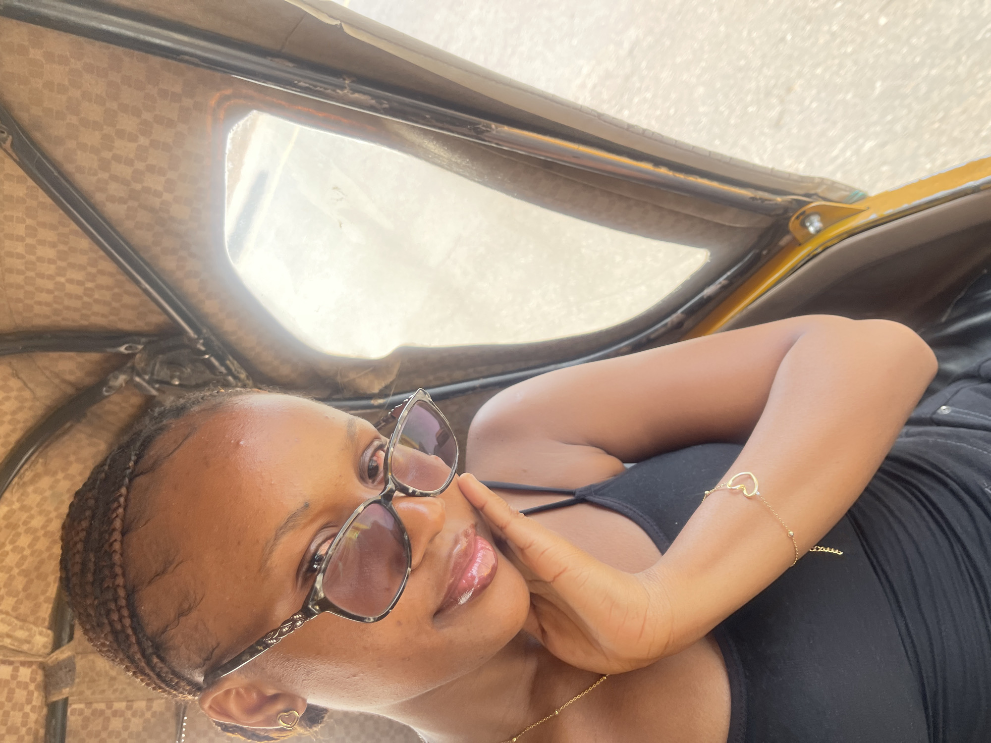 A young woman with braided hair and glasses is taking a selfie inside a rickshaw. She is wearing a black tank top and a gold bracelet with a heart charm. The interior of the rickshaw is lined with a brown checkered fabric. The scene appears to be in Rafin Par, Nigeria, judging by the context provided, although this cannot be definitively confirmed from the image alone.