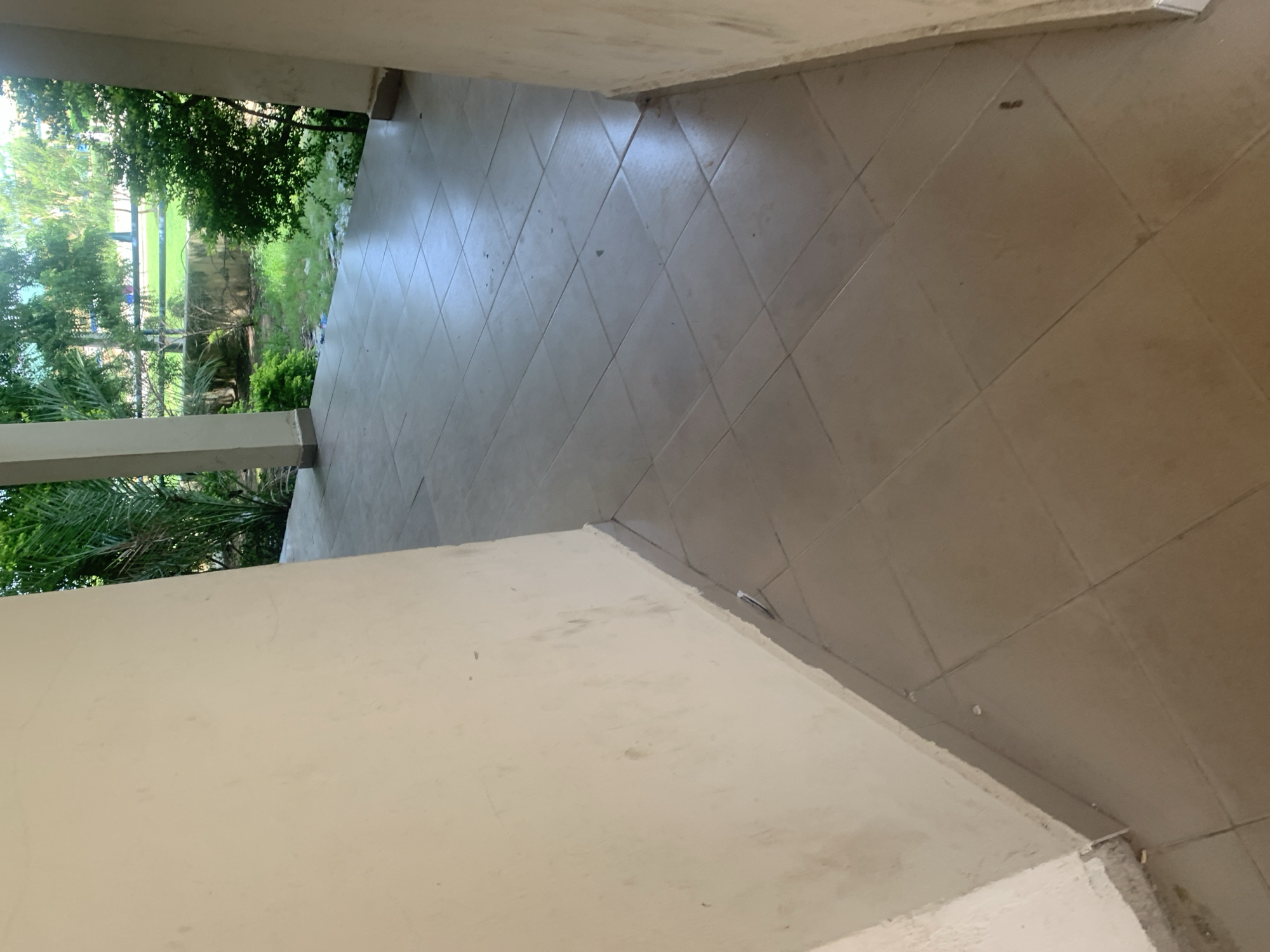 The media file depicts a tiled walkway or corridor. The tiles are laid in a diamond pattern and are a light, neutral color. The walkway is bordered on one side by a light-colored wall or pillar. On the other side, there is an opening that looks out onto a lush green outdoor space with trees and foliage. The outdoor area appears to be well-lit, suggesting it is daytime. The overall scene conveys a sense of an outdoor or semi-outdoor passageway, possibly at a building or facility. There is no visible text in the media file. The location context provided is Jalingo, Nigeria.