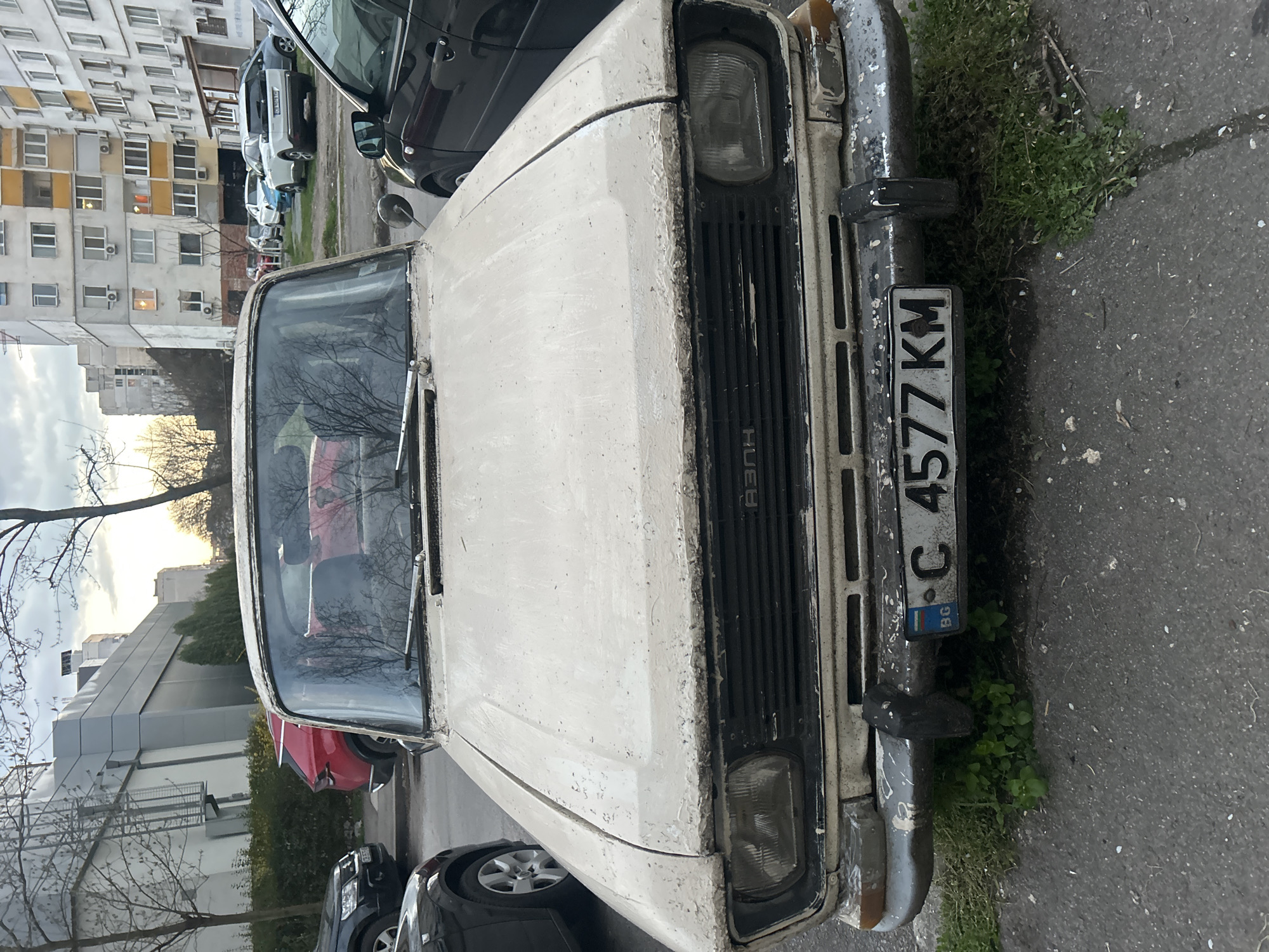The image shows the front of a weathered, light-colored car, likely an older model, parked on a street. The license plate, "C 4577 KM," is affixed to the front bumper. The car appears to be located in an urban area, evidenced by the presence of buildings with numerous windows in the background. Other parked cars are also visible on the street. Grass and small plants are growing near the bumper, suggesting a lack of recent movement or maintenance of the area. The sky is overcast, hinting at the time of day being either early morning or late afternoon.