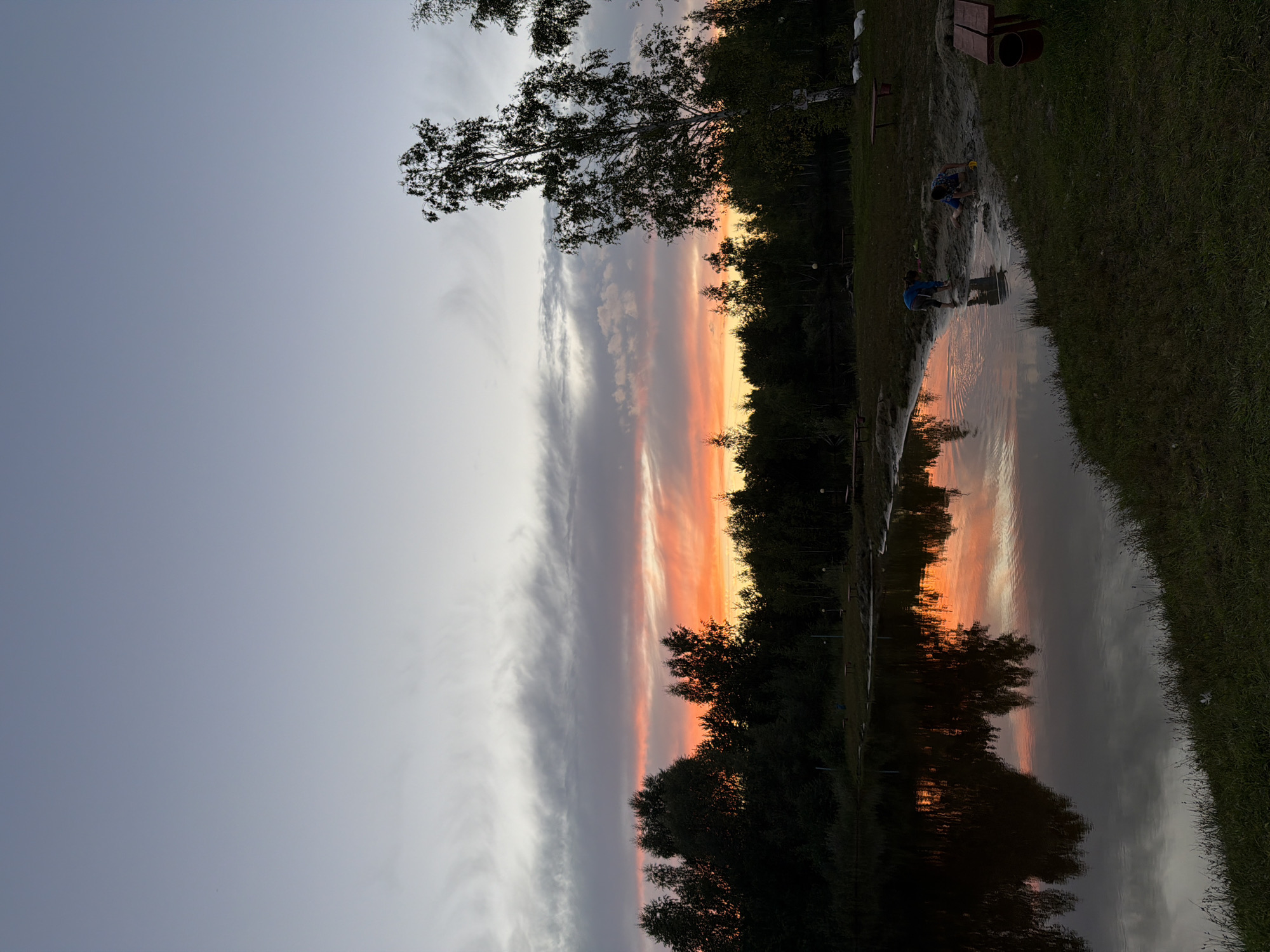 The image captures a serene sunset scene at a lake located in the vicinity of Glubokovo, Russia. The sky is painted with hues of soft grey and pastel orange, with elongated clouds stretching across the horizon, reflecting vibrantly on the calm water. A dense line of dark green trees forms a natural border along the far bank of the lake, silhouetted against the colorful sky. In the foreground, the grassy bank slopes gently towards the water. On the sandy edge of the lake, two young individuals, appearing to be boys, are engaged in playing with sand. One is standing, partially in the water, while the other is crouched down, actively digging. Scattered around the sandy area are a few wooden benches and a red trash can, suggesting a recreational area. Tall trees, one of which is a prominent birch with its distinctive white bark, stand on the right side of the frame, adding to the natural landscape. The overall impression is one of a peaceful evening by the water, marked by the play of children.