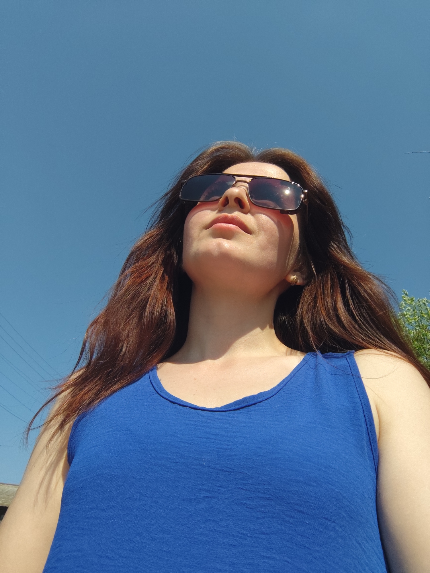A woman with long, auburn hair is looking upwards, wearing sunglasses and a blue tank top. The sky is clear and blue. Power lines are visible in the background. Green foliage is present on the right side of the frame. The location cannot be determined to be Фоки, Russia.