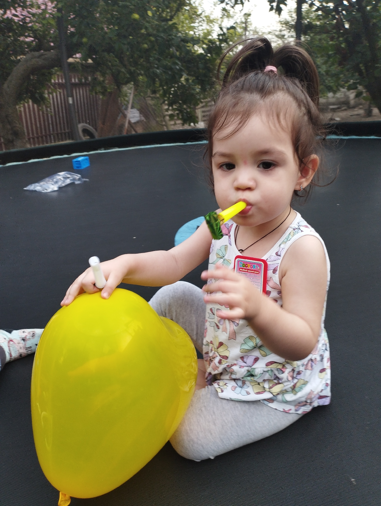 The image depicts a young child on a trampoline. The child, a girl with dark hair styled in two pigtails, is wearing a white sleeveless top with a colorful bow pattern and grey leggings. She is holding a yellow balloon in her left hand and blowing into a yellow party blower with a green frill in her mouth. The trampoline surface is black. In the background, trees and a wooden fence are visible, suggesting an outdoor setting, possibly a backyard. A small blue plastic object and a crumpled foil wrapper are also present on the trampoline. There are no indications of the image being specifically located in the city of Fălești, Moldova.