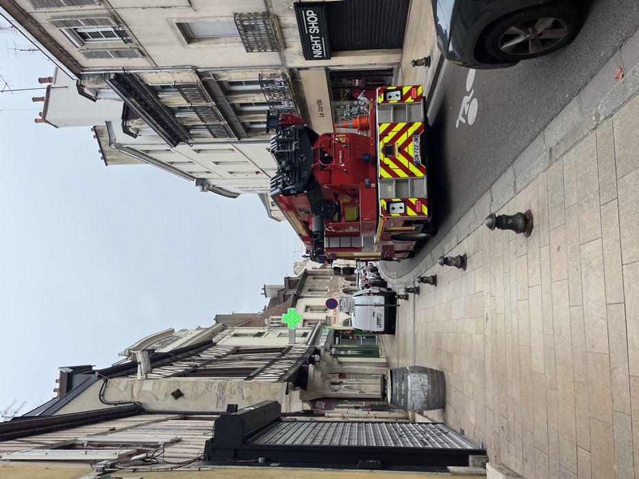 This image depicts a narrow historic street in Dijon, France, under an overcast sky. A large, red fire engine with an elevated articulated ladder/boom system occupies the center of the asphalt street, facing away from the viewer. Its rear displays prominent yellow and red chevron reflective markings and a French license plate, GN-722-JM. Two orange traffic cones are placed behind the fire engine on the right.
On the left, a wide sidewalk paved with light-colored rectangular flagstones features a weathered wooden barrel with metal hoops in the foreground. Several black, ornate bollards line the curb. Buildings on the left side include a multi-story structure with exposed dark timber beams and light infill, characteristic of half-timbered architecture. Further down, an illuminated green cross sign marks a pharmacy, near which a white commercial van is parked. Other vehicles are visible further down the street, which curves gently to the left.
On the right side of the street, multi-story buildings with light-colored facades are visible. One building features dark metal balconies and windows with light-colored shutters. A shop entrance below is labeled "Le Cercle." Adjacent to it, a storefront with a closed metallic roller shutter displays "NIGHT SHOP." The front portion of a dark-colored vehicle is parked in the extreme foreground on the right. A white bicycle lane symbol is painted on the asphalt near the right curb. Overhead wires and antennas are visible on the rooftops. No people are present in the scene.