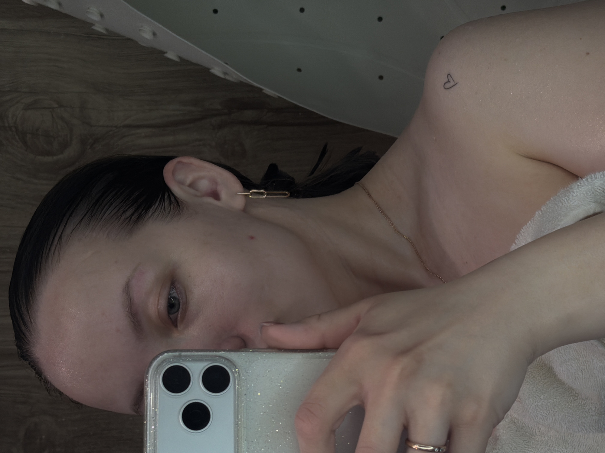 A selfie taken by a fair-skinned person with wet, dark hair. The individual has light eyes and wears a gold chain necklace and a gold earring shaped like a paperclip link. A small heart tattoo is visible on the upper left shoulder. The person is holding a glittery white smartphone with three camera lenses. Part of a white towel is visible. The background includes a wooden-patterned surface and a white object with small black dots. The location of Reshetnikovo, Russia, cannot be determined from the image.